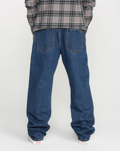 A person wearing a plaid shirt and Volcom Men's Pipe Dreams Denim Pants in a dark indigo wash stands facing away, showcasing the back of the jeans and white sneakers against a plain background.