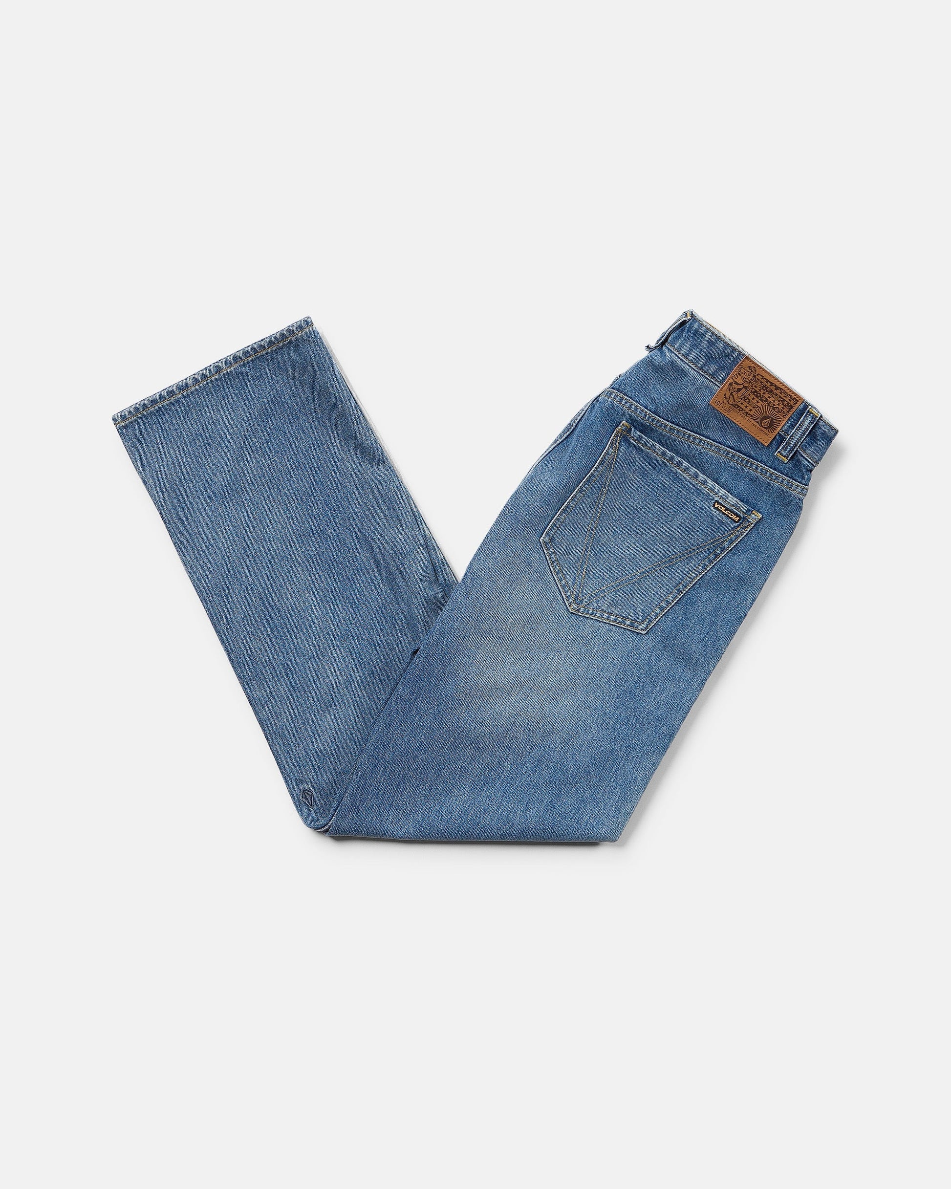A pair of Volcom Men's Pipe Dreams Denim Pants in Euro Light Blue is folded with one leg crossed under the other, revealing a back pocket and a brown leather patch on the waistband, set against a white background.