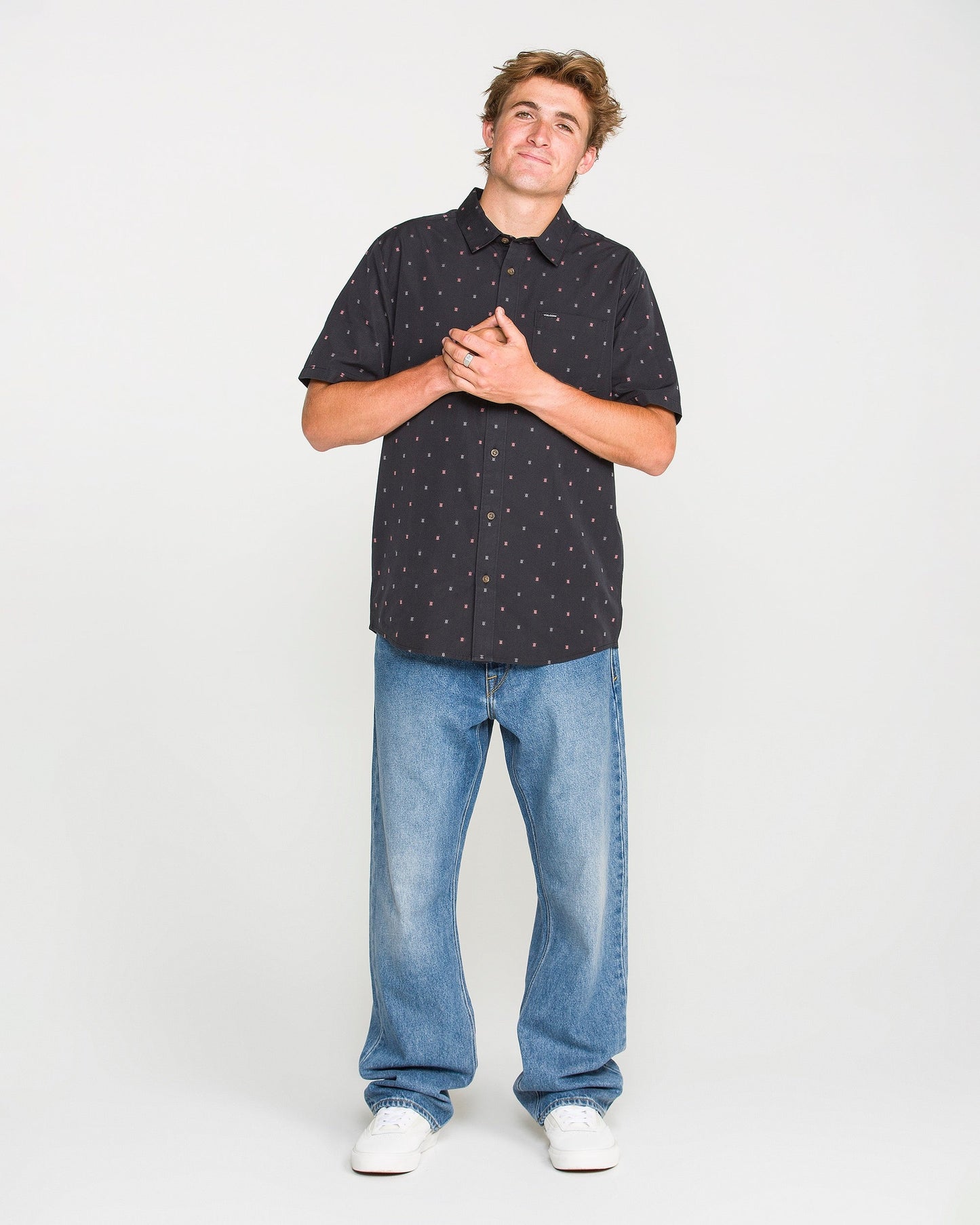 A man stands against a white background, wearing a black short-sleeved button-up shirt with a small pattern, Volcom's Men's Pipe Dreams Denim Pants in a vintage-inspired relaxed straight fit, and white sneakers. He clasps his hands over his chest and looks slightly upwards.