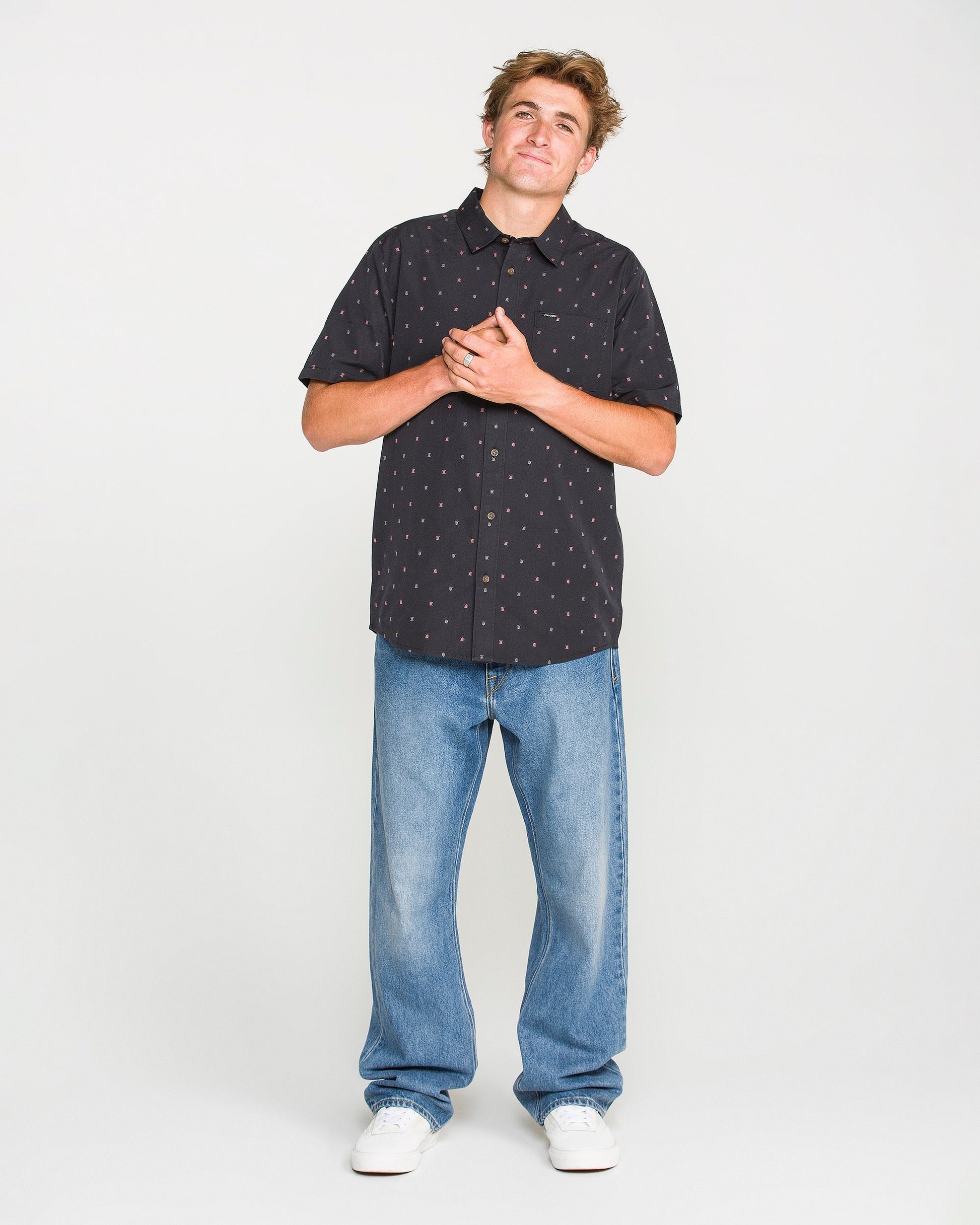 A man stands against a white background, wearing a black short-sleeved button-up shirt with a small pattern, Volcom's Men's Pipe Dreams Denim Pants in a vintage-inspired relaxed straight fit, and white sneakers. He clasps his hands over his chest and looks slightly upwards.