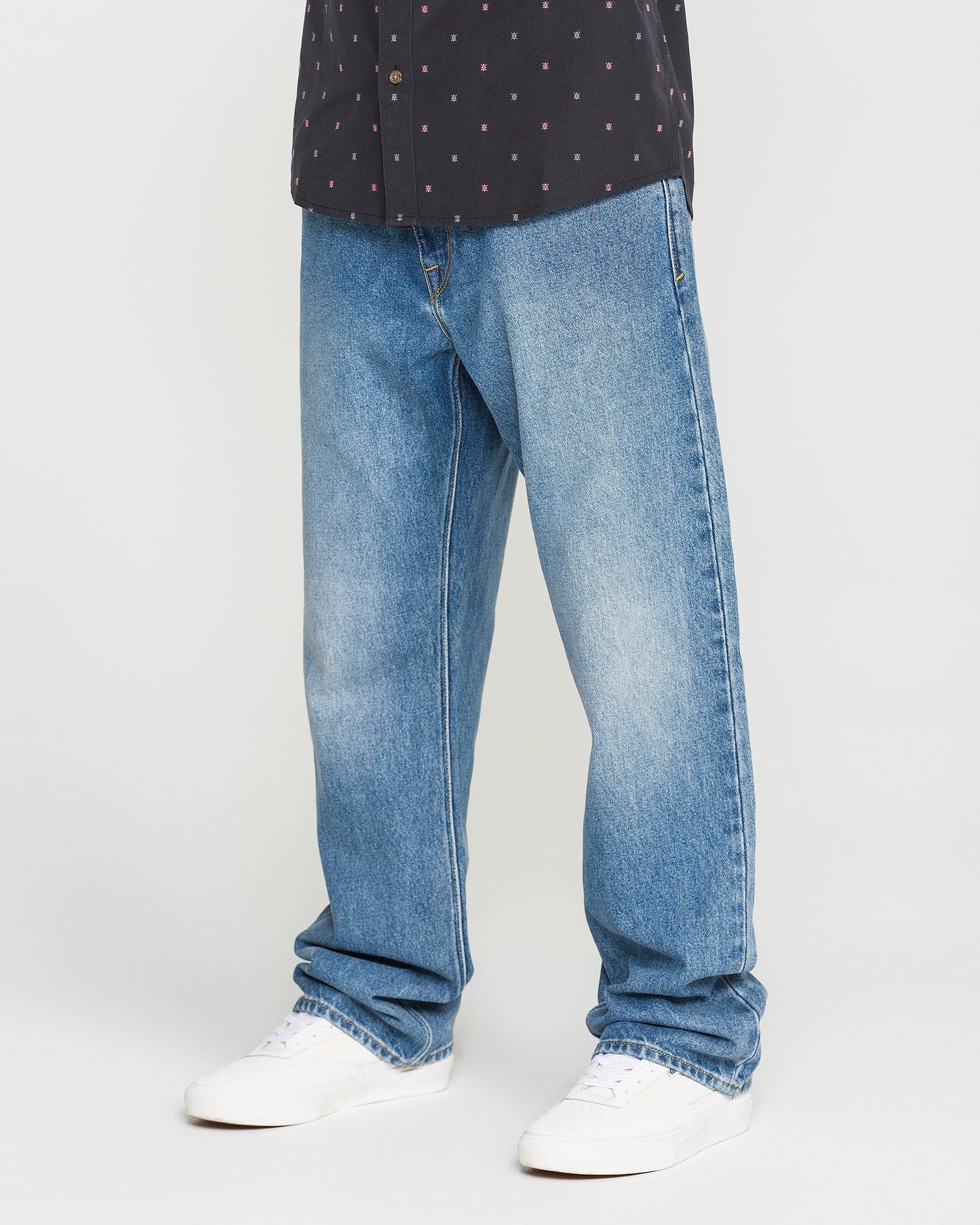 A person is shown from the waist down wearing Volcom Men's Pipe Dreams Denim Pants in Euro Light Blue, styled with white sneakers and a black patterned shirt, standing against a plain background.