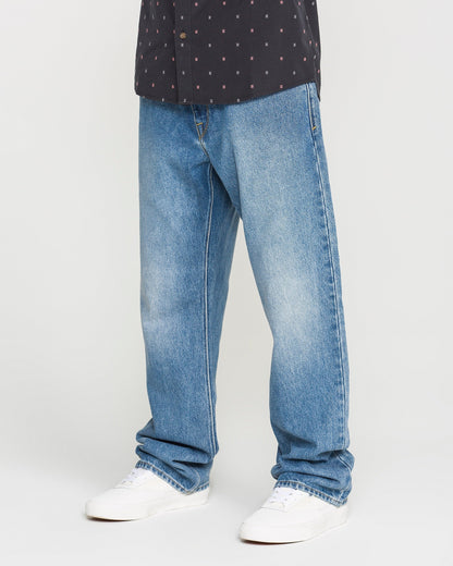 A person is shown from the waist down wearing Volcom Men's Pipe Dreams Denim Pants in Euro Light Blue, styled with white sneakers and a black patterned shirt, standing against a plain background.
