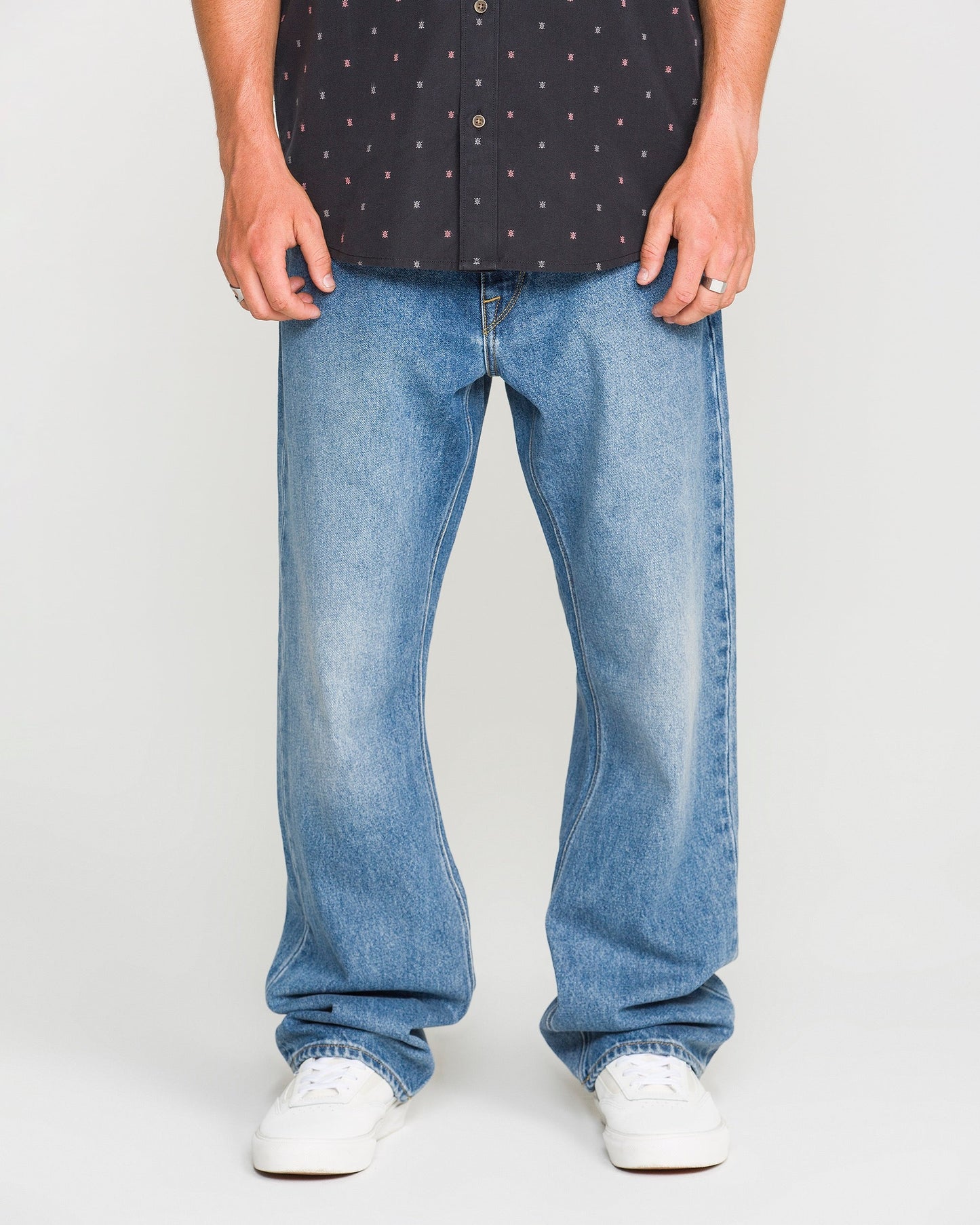 A person wearing a black patterned shirt, Volcom Men's Pipe Dreams Denim Pants in a relaxed straight fit, and white sneakers stands against a plain background. Only their legs and hands are visible.