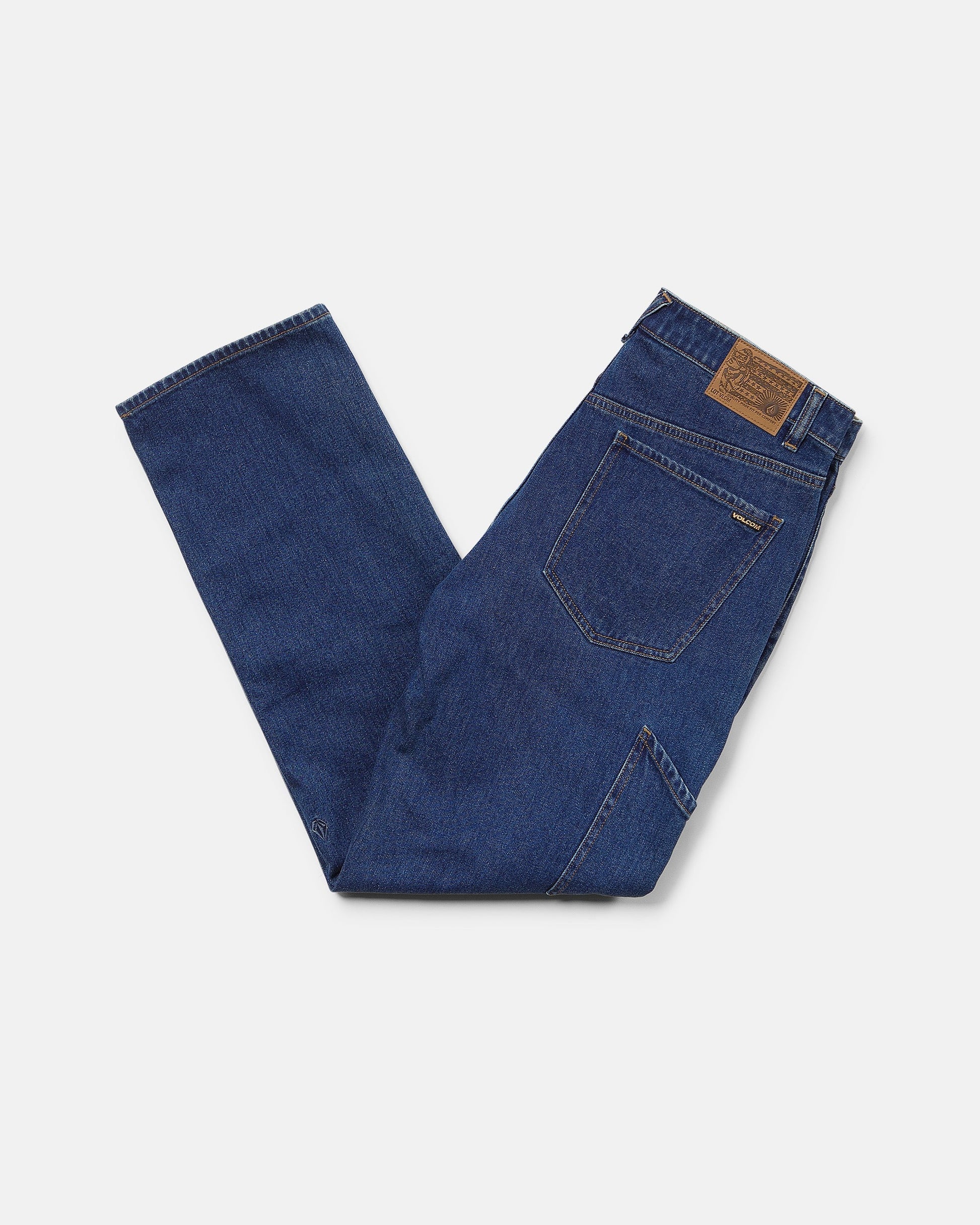 A pair of folded Volcom Men's Kinkade Utility Denim Pants in indigo, featuring visible back pockets and a brown leather patch on the waistband, displayed on a white background.