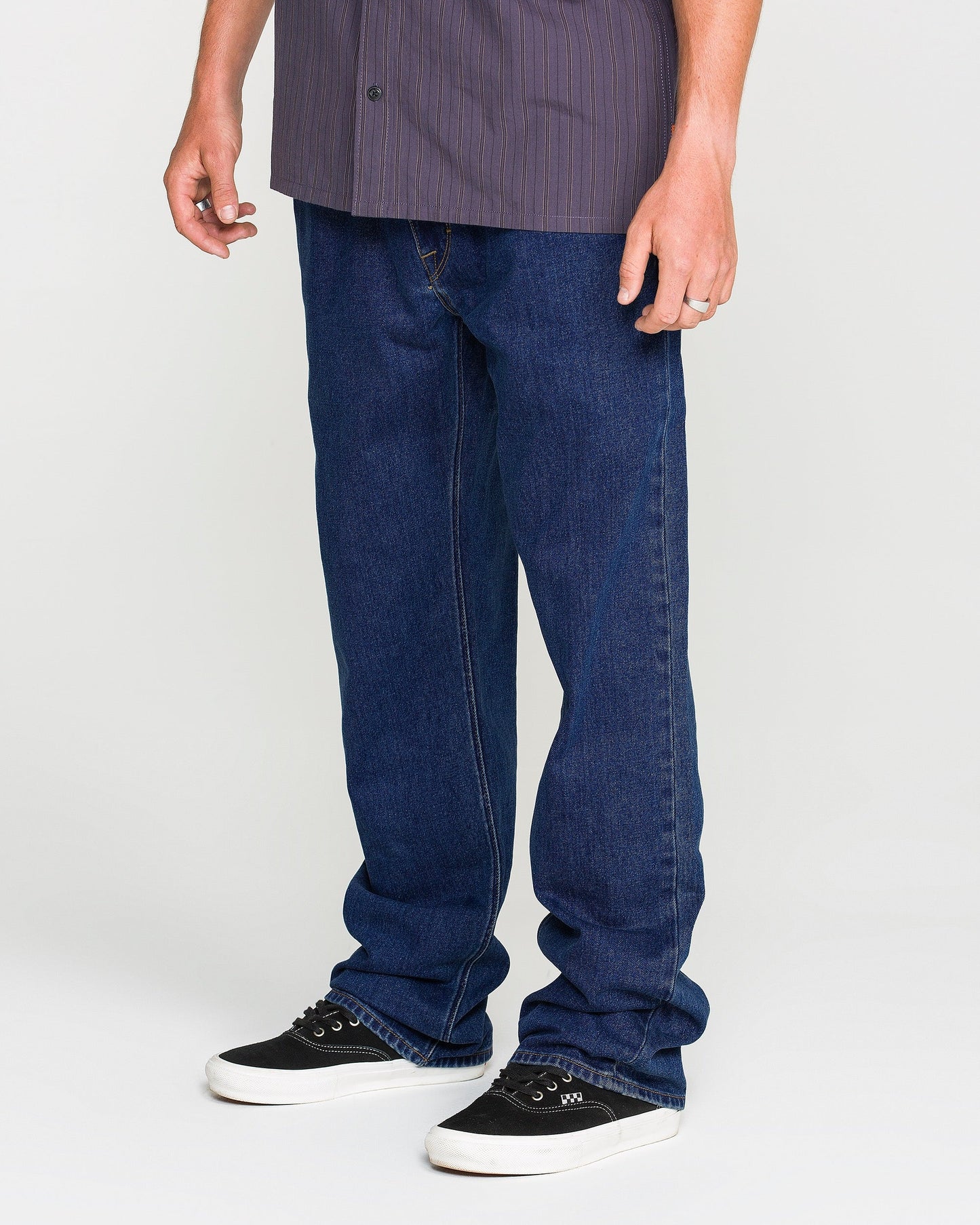 A person wearing a short-sleeved, dark striped shirt, Volcom Men's Kinkade Utility Denim Pants in an indigo wash, and black sneakers with white soles stands against a plain white background.