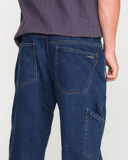 A person wearing Volcom Men's Kinkade Utility Denim Pants in dark blue indigo, featuring visible back and side pockets, paired with a purple striped shirt. The photo is taken from behind, highlighting the straight-leg utility denim pants and part of the shirt.