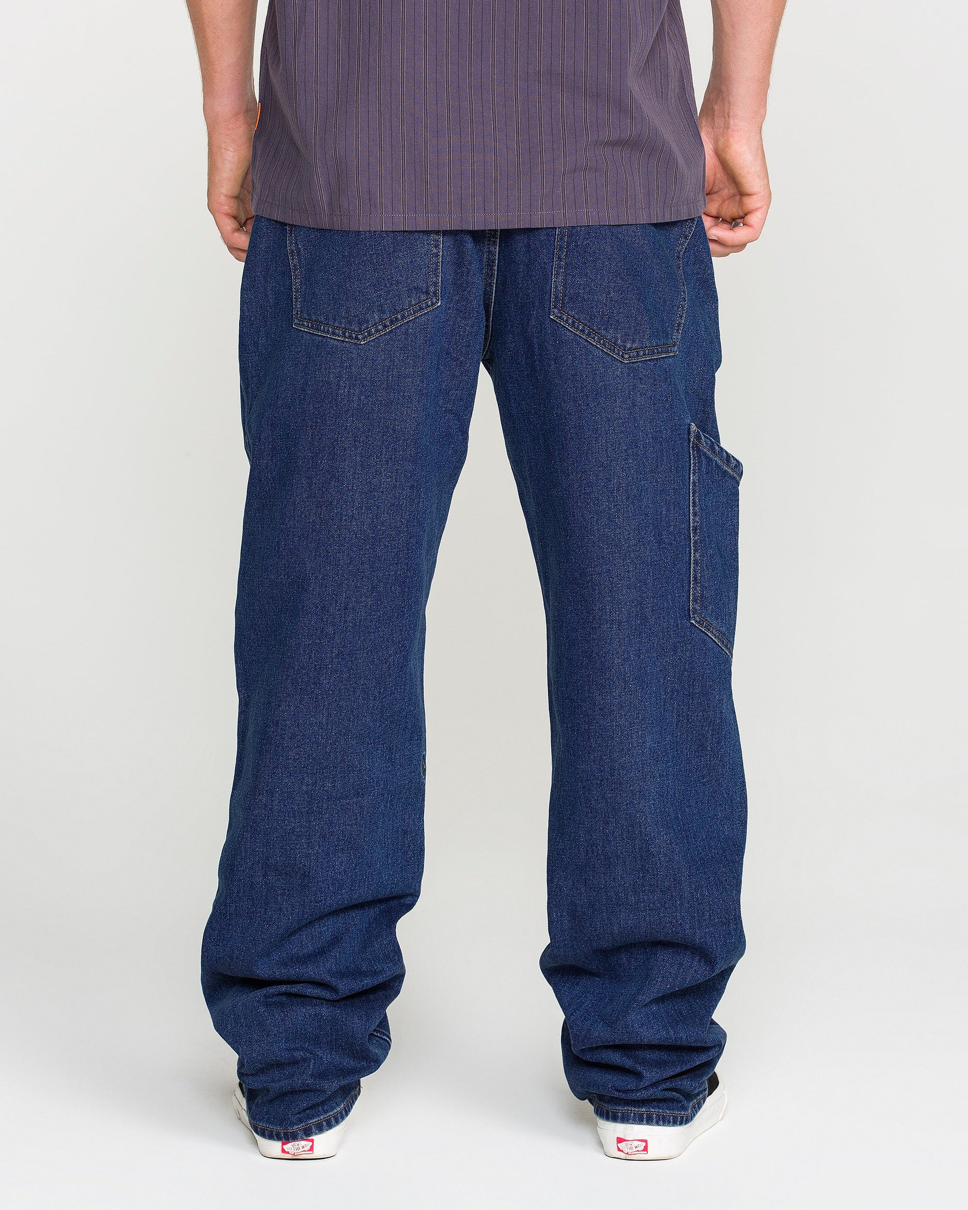 A person wearing Volcom Men's Kinkade Utility Denim Pants in indigo and white sneakers is standing facing away from the camera, with only their lower back, legs, and feet visible.