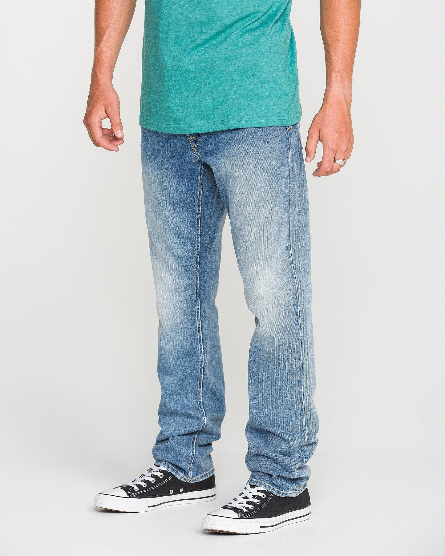 A person wearing a green t-shirt, Volcom Men's Vorta Denim Pants in a slim fit, and black Converse sneakers stands against a plain white background. Their face is not visible.