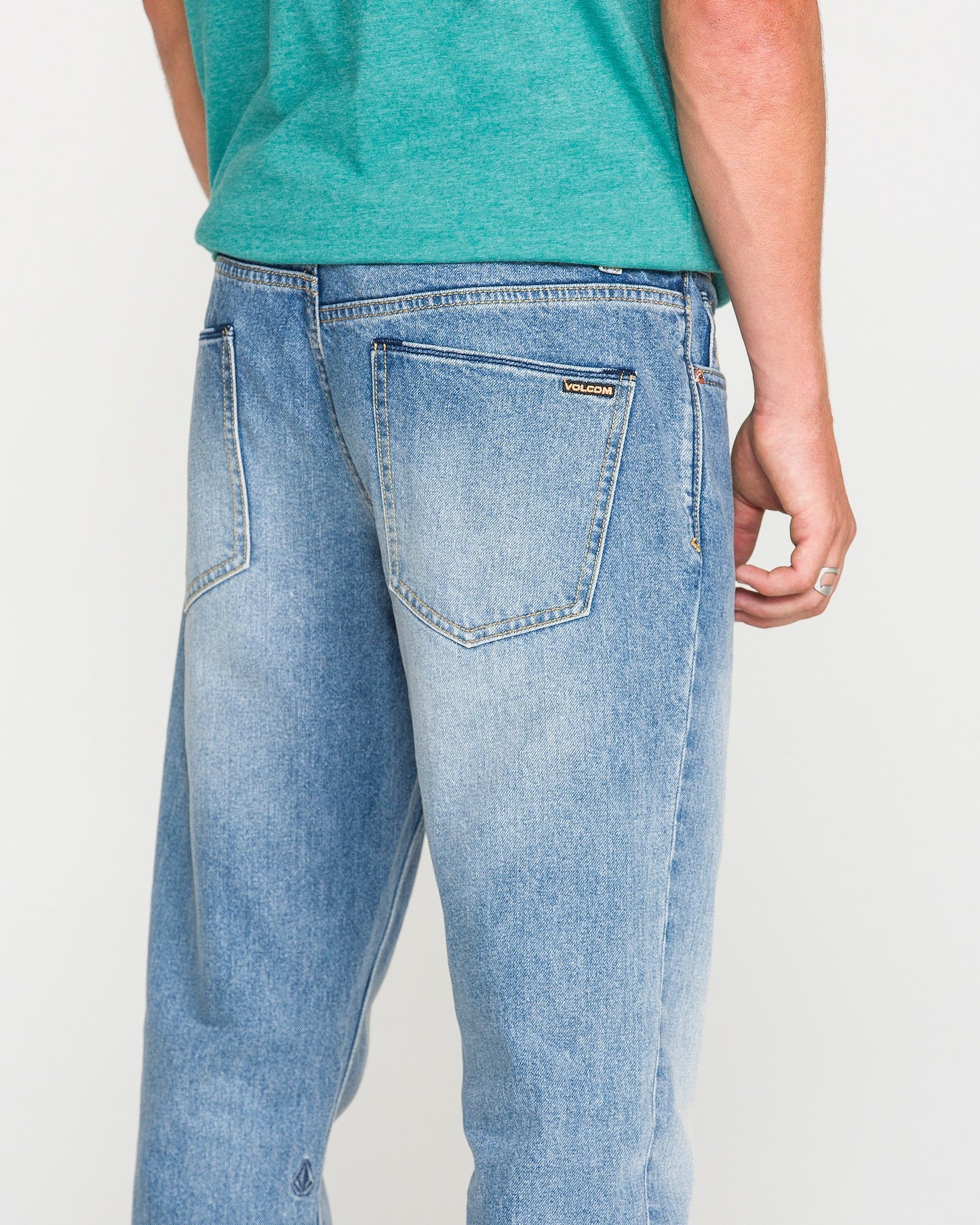 A person in a green shirt and light blue Volcom Men's Vorta Denim Pants is shown from the back, with one arm relaxed at their side. The Vorta denim features two back pockets and a small brand label above the right pocket.