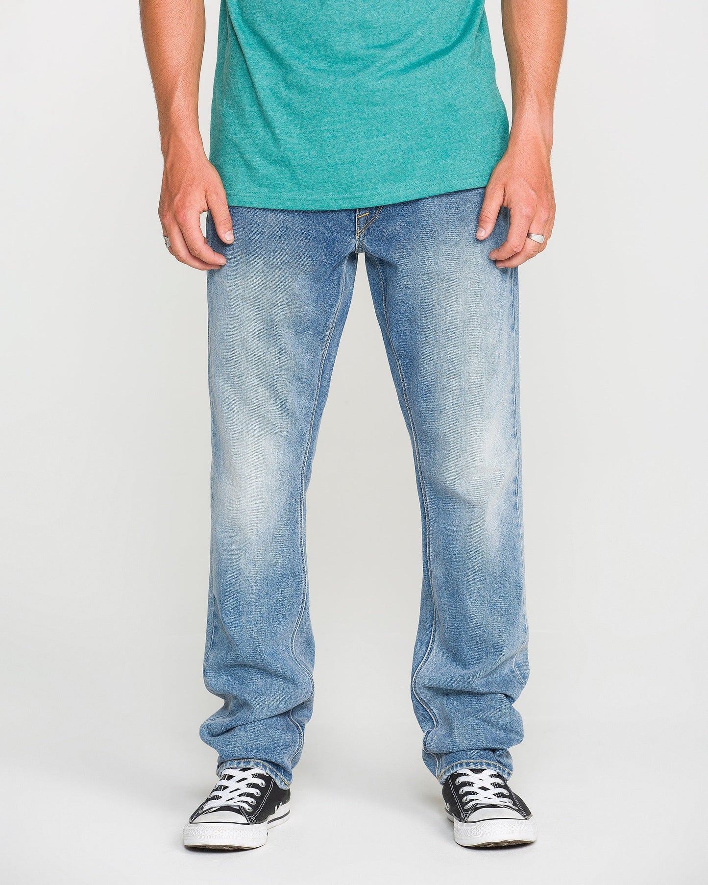 A person wearing a green t-shirt, Volcom Men's Vorta Denim Pants in a slim fit, and black-and-white sneakers stands with arms relaxed at their sides against a plain background.