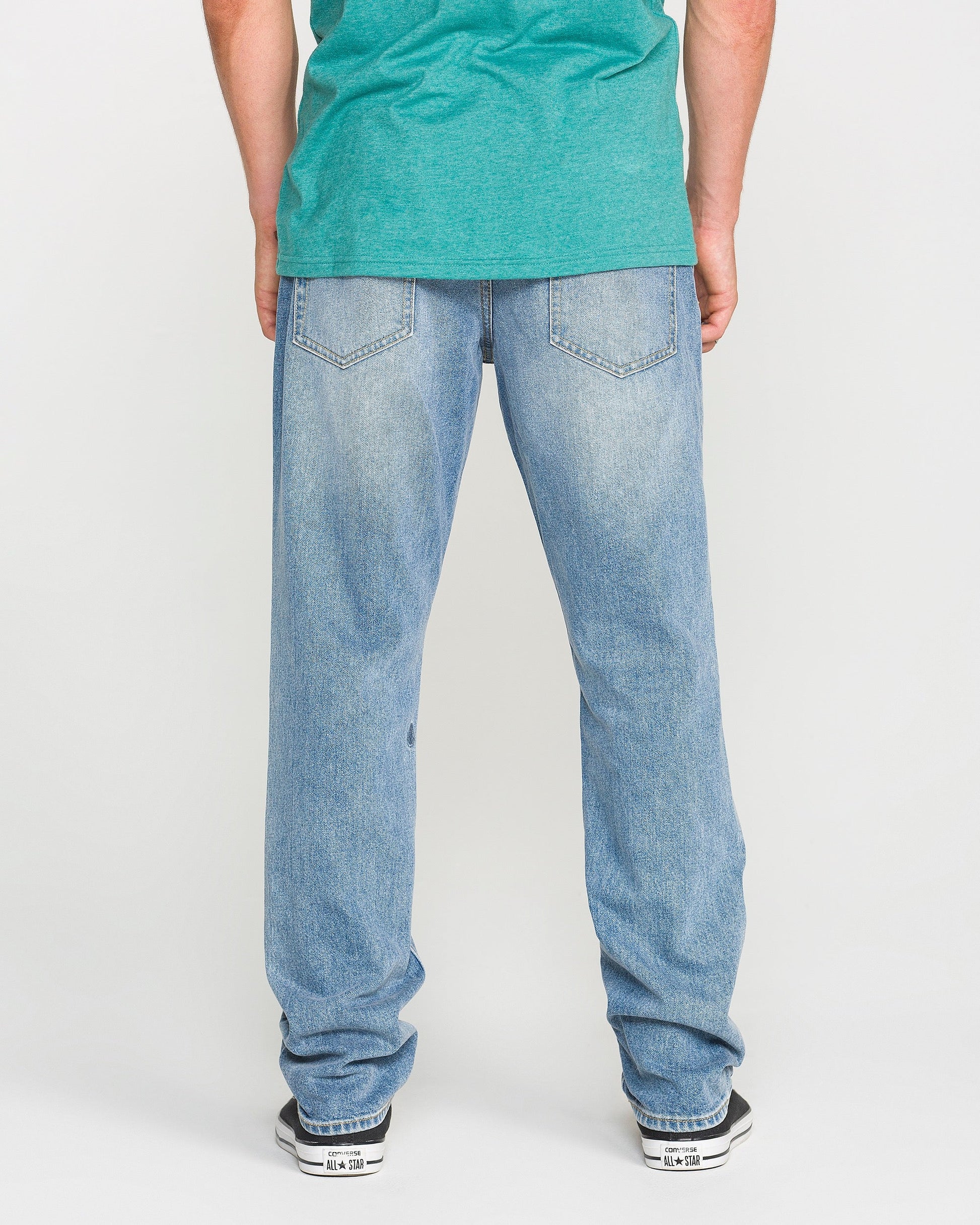 A person wearing a green T-shirt, Volcom Men's Vorta Denim Pants in Freedom Blue (slim fit), and black-and-white sneakers stands facing away, showcasing the back view of the jeans.