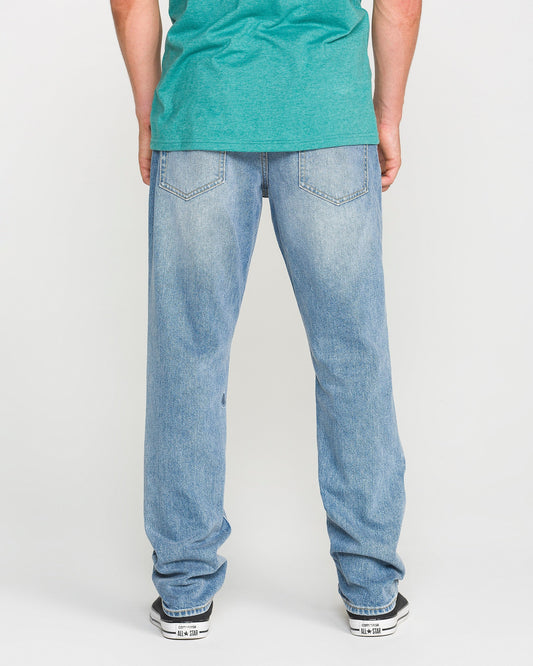 A person wearing a green T-shirt, Volcom Men's Vorta Denim Pants in Freedom Blue (slim fit), and black-and-white sneakers stands facing away, showcasing the back view of the jeans.