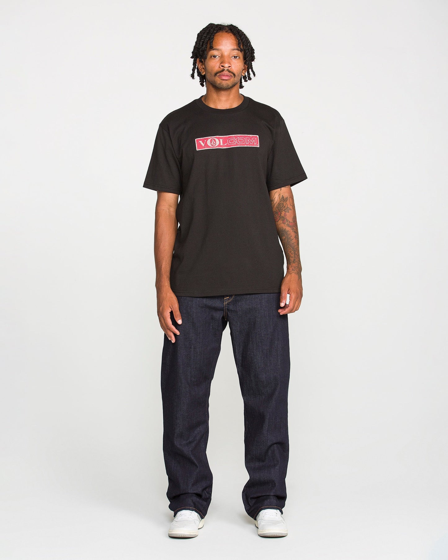 A person with short dreadlocks wears a black Volcom t-shirt, Men's Pipe Dreams Denim Pants by Volcom in indigo stretch denim featuring signature details, and white sneakers, standing against a plain white background.