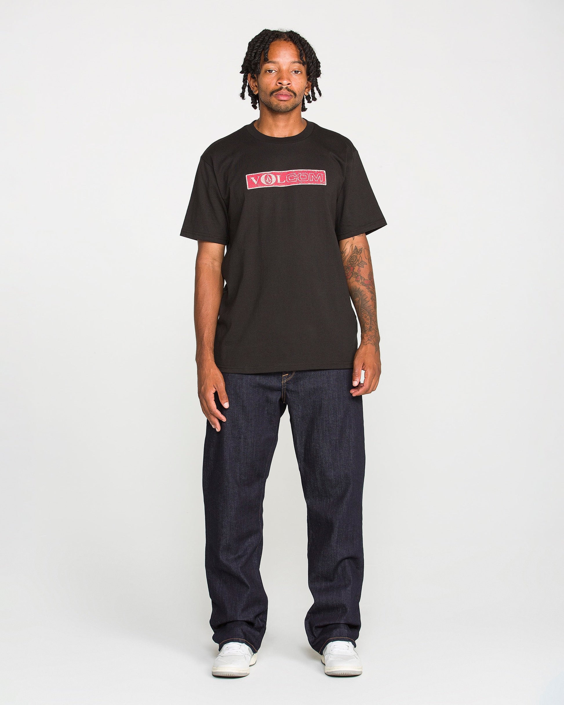 A person with short dreadlocks wears a black Volcom t-shirt, Men's Pipe Dreams Denim Pants by Volcom in indigo stretch denim featuring signature details, and white sneakers, standing against a plain white background.
