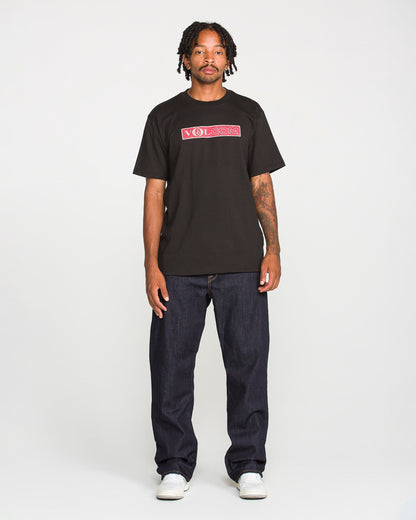 A person with short dreadlocks wears a black Volcom t-shirt, Men's Pipe Dreams Denim Pants by Volcom in indigo stretch denim featuring signature details, and white sneakers, standing against a plain white background.