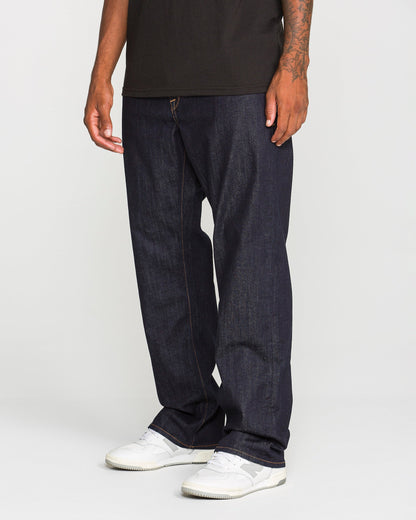A person wearing a black t-shirt, white sneakers, and Volcom Men's Pipe Dreams Denim Pants—relaxed straight-leg jeans crafted from indigo stretch denim—stands against a plain background. Only the lower body and one tattooed arm are visible, highlighting subtle Volcom signature details for an effortlessly cool look.