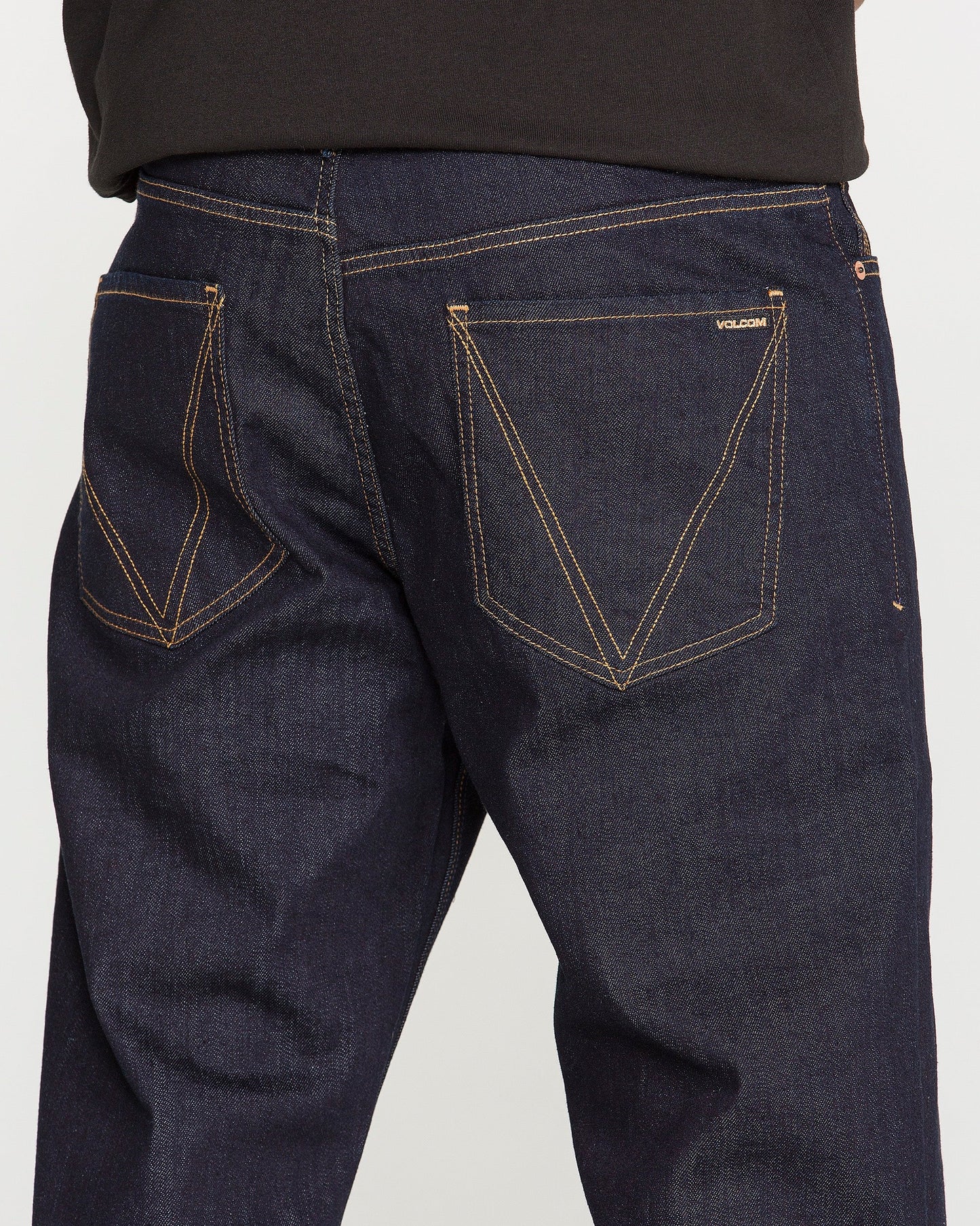 Close-up of the back of a person wearing Volcom Men's Pipe Dreams Denim Pants in dark blue indigo stretch denim, featuring contrasting orange stitching and two back pockets—one adorned with a small “Volcom” label that showcases the brand's signature details.
