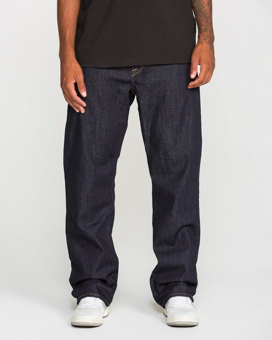 A person stands against a plain background, wearing Volcom Men's Pipe Dreams Denim Pants in indigo stretch denim with a relaxed straight leg fit and signature brand details, paired with white sneakers and a black t-shirt. Only their lower torso and legs are visible.