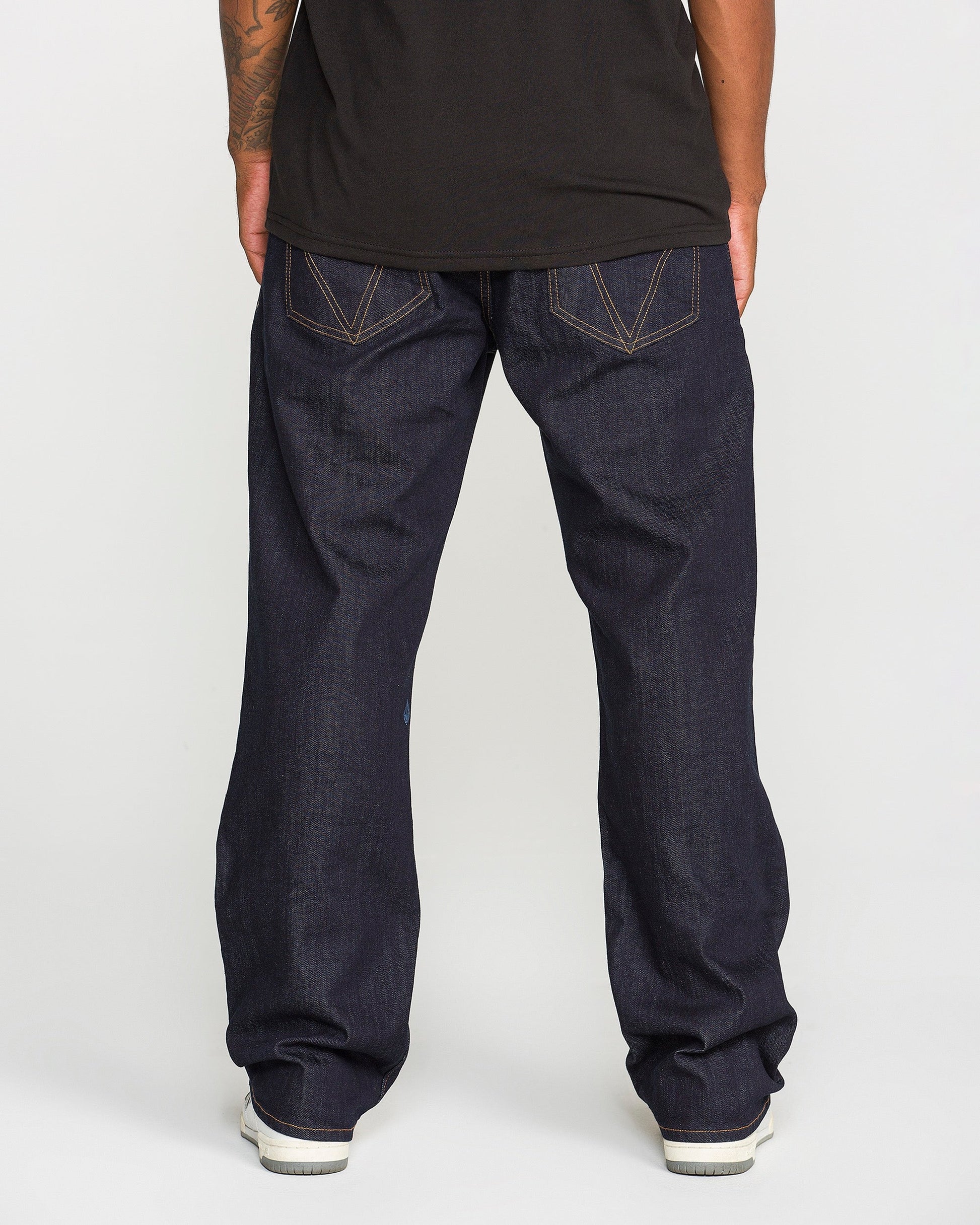 A person, shown from the waist down, wears Volcom Men's Pipe Dreams Denim Pants in a dark blue indigo wash with visible stitching and signature branding. The relaxed straight leg jeans are paired with a black shirt and white sneakers.