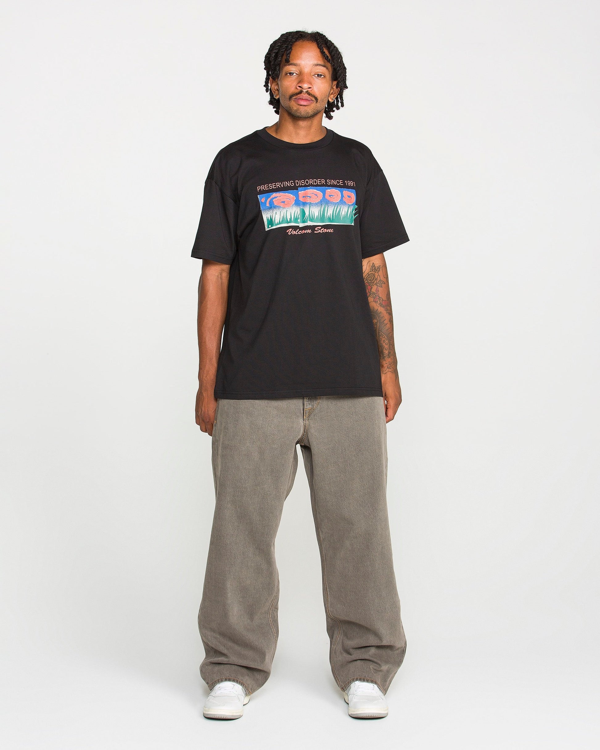 A man with short dreadlocks stands against a plain white background wearing a black graphic T-shirt, white sneakers, and Volcom Men's Featured Artist Antonio Carrau Reverb Pants in loose gray overdyed black cotton.