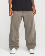 A person stands against a plain white background, cropped at the shoulders, wearing Volcom Men's Featured Artist Antonio Carrau Reverb Pants in gray, paired with a black overdyed cotton T-shirt and white sneakers.