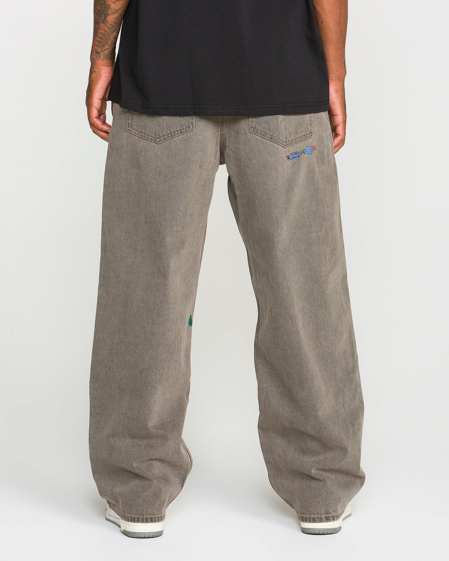 A person is standing with their back to the camera, wearing Volcom Men’s Featured Artist Antonio Carrau Reverb Pants—loose-fitting, low-rise gray jeans with small embroidered details on the back pockets—and a black shirt made of overdyed cotton. White shoes are partially visible.