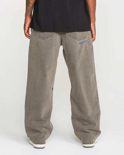 A person is standing with their back to the camera, wearing Volcom Men’s Featured Artist Antonio Carrau Reverb Pants—loose-fitting, low-rise gray jeans with small embroidered details on the back pockets—and a black shirt made of overdyed cotton. White shoes are partially visible.