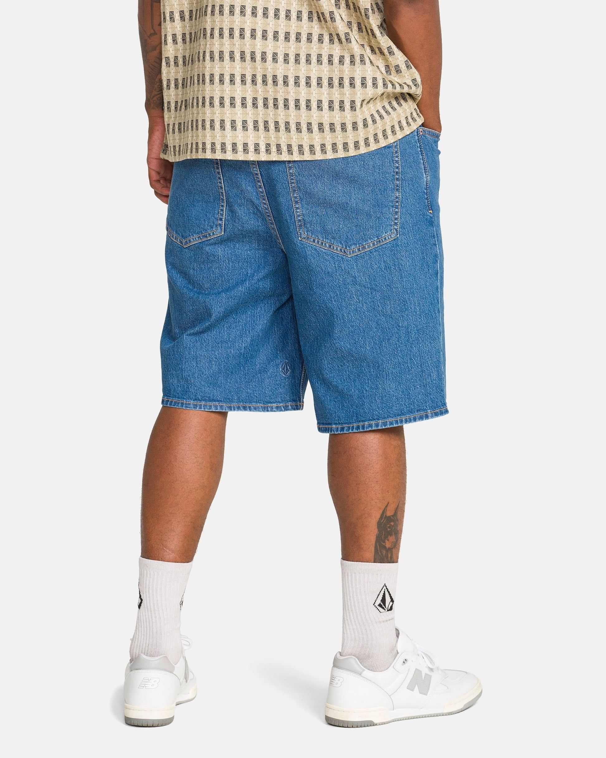 A person stands facing away, wearing Volcom Men's Billow Denim Shorts, a beige patterned shirt, white crew socks with a black logo, and white New Balance sneakers. A tattoo is visible on their left calf.