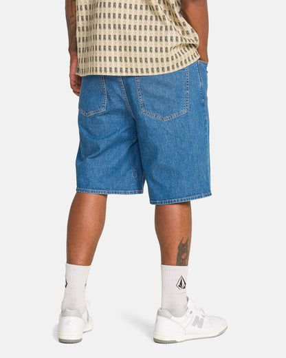 A person stands facing away, wearing Volcom Men's Billow Denim Shorts, a beige patterned shirt, white crew socks with a black logo, and white New Balance sneakers. A tattoo is visible on their left calf.