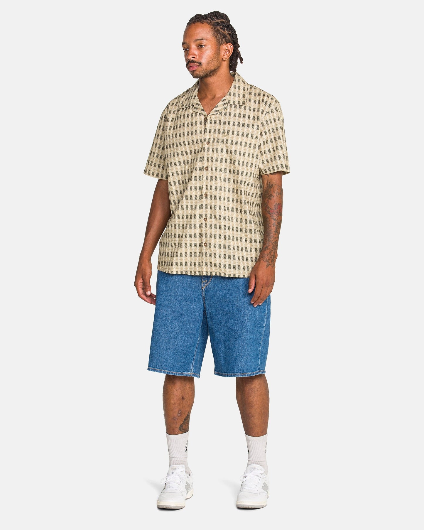 A man with tattoos and dreadlocks wears a beige patterned short-sleeve shirt, Volcom Men's Billow Denim Shorts, white socks, and white sneakers while standing against a plain white background.
