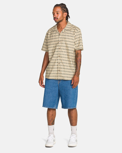 A man with tattoos and dreadlocks wears a beige patterned short-sleeve shirt, Volcom Men's Billow Denim Shorts, white socks, and white sneakers while standing against a plain white background.