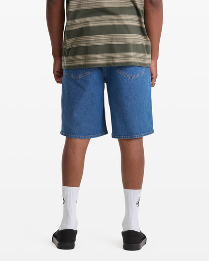 A person stands facing away from the camera against a white background wearing a green striped shirt, Men's Billow Denim Short 22 by Volcom, white socks, and black slide sandals.