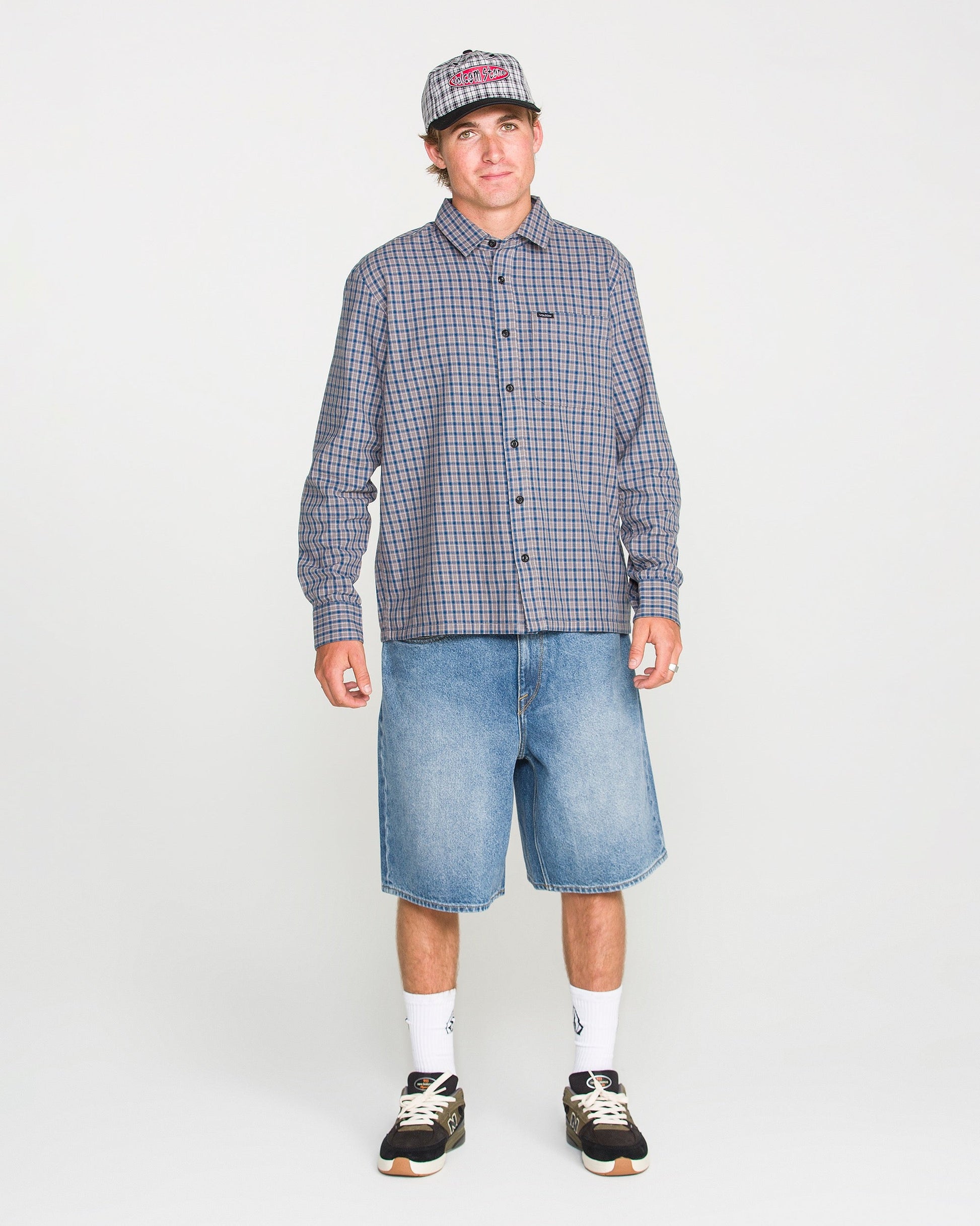 A person wearing a loose fit plaid shirt, Volcom Men's Billow Denim Shorts, sneakers, white socks, and a baseball cap stands against a plain white background.