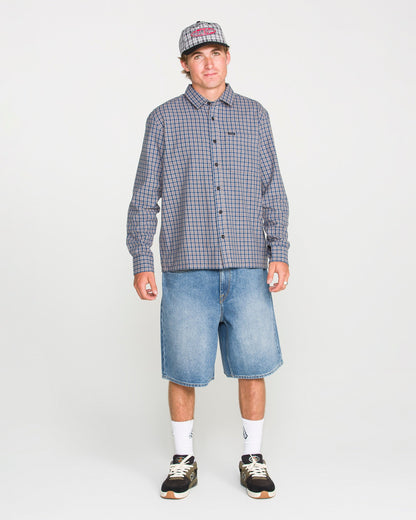 A person wearing a loose fit plaid shirt, Volcom Men's Billow Denim Shorts, sneakers, white socks, and a baseball cap stands against a plain white background.