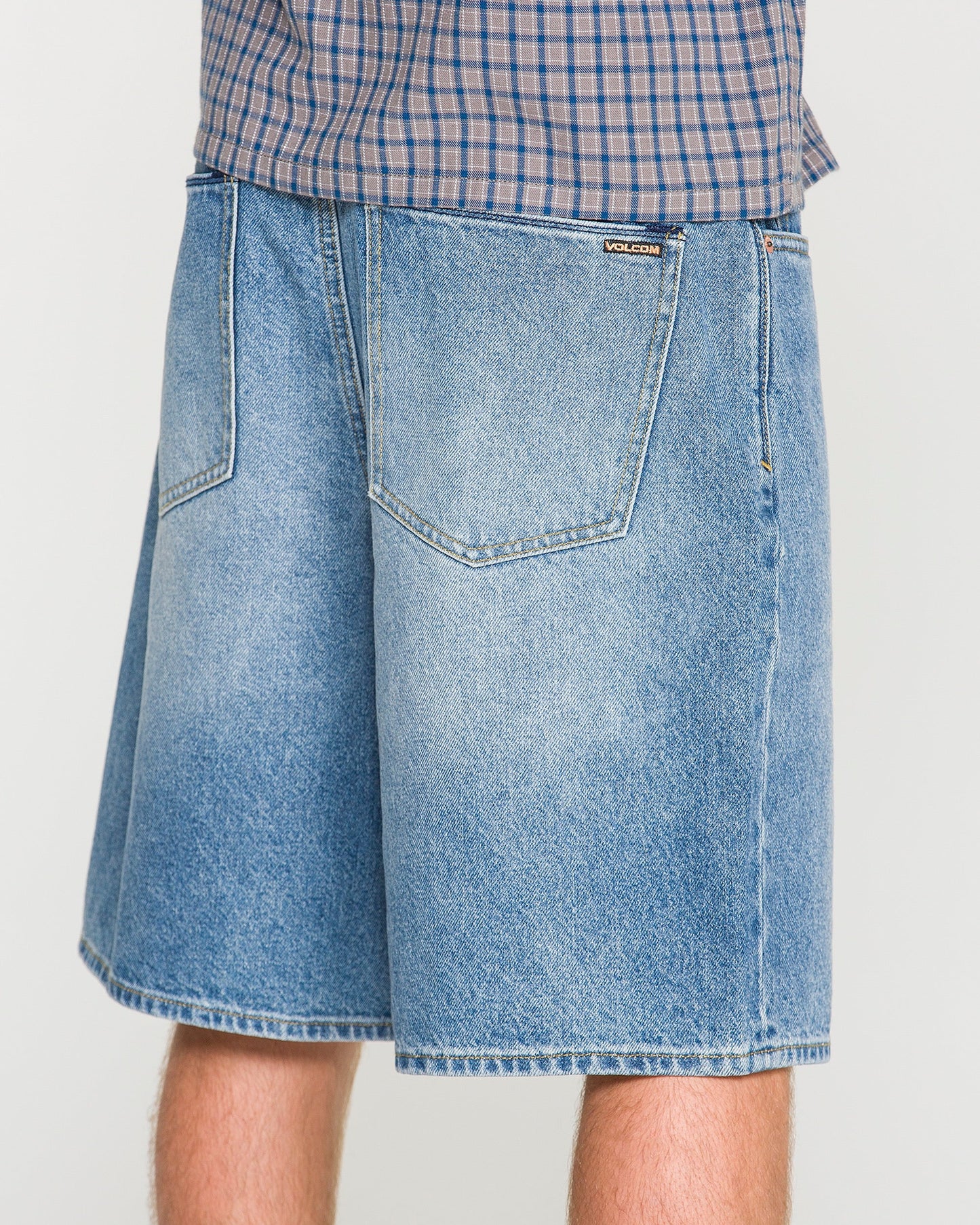 A person wearing loose fit, light blue Men's Billow Denim Shorts by Volcom and a plaid shirt, shown from the waist down, facing away from the camera.
