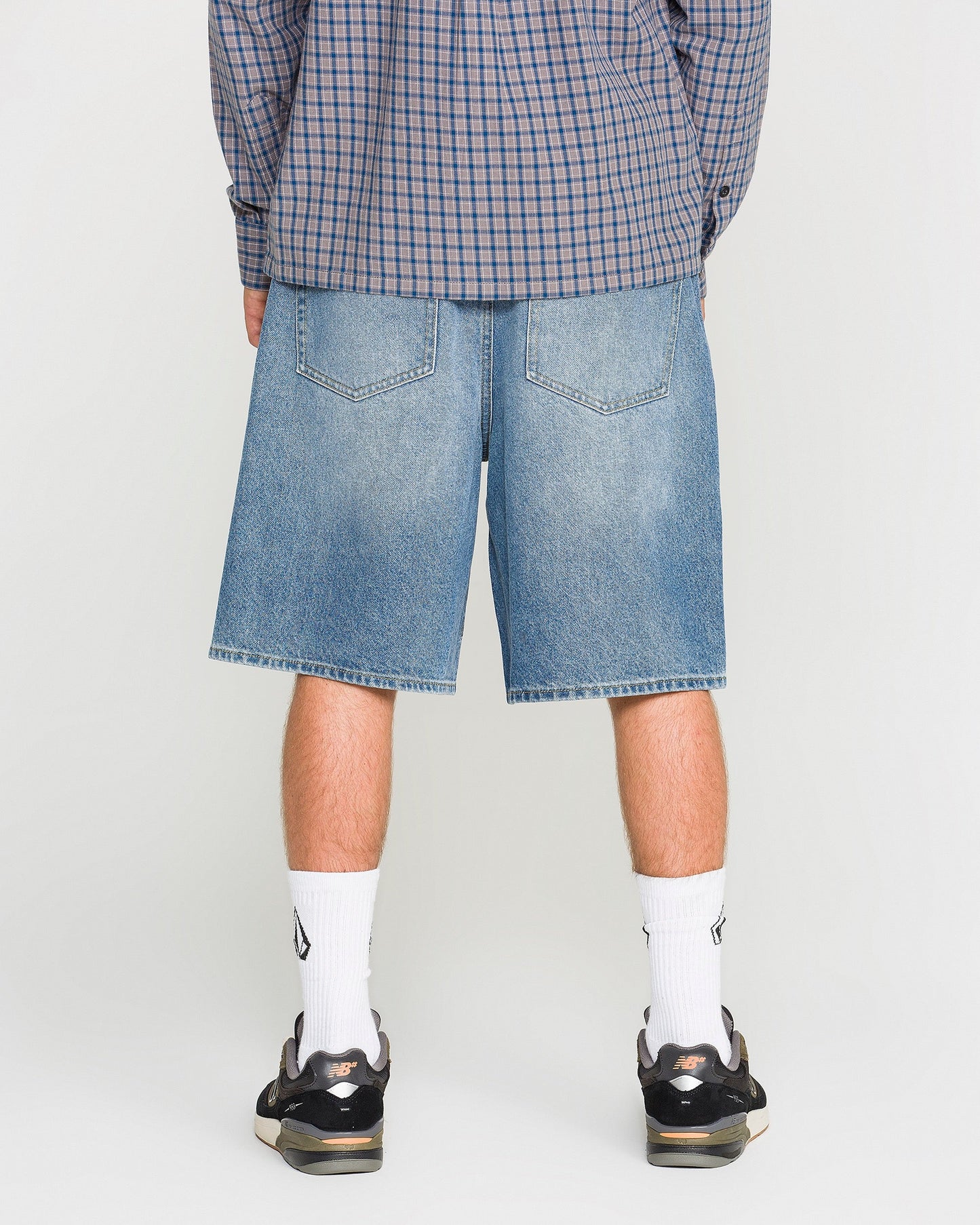 A person wearing a blue plaid shirt, Volcom Men's Billow Denim Shorts in a loose fit, white socks, and black sneakers is standing with their back to the camera against a plain background.