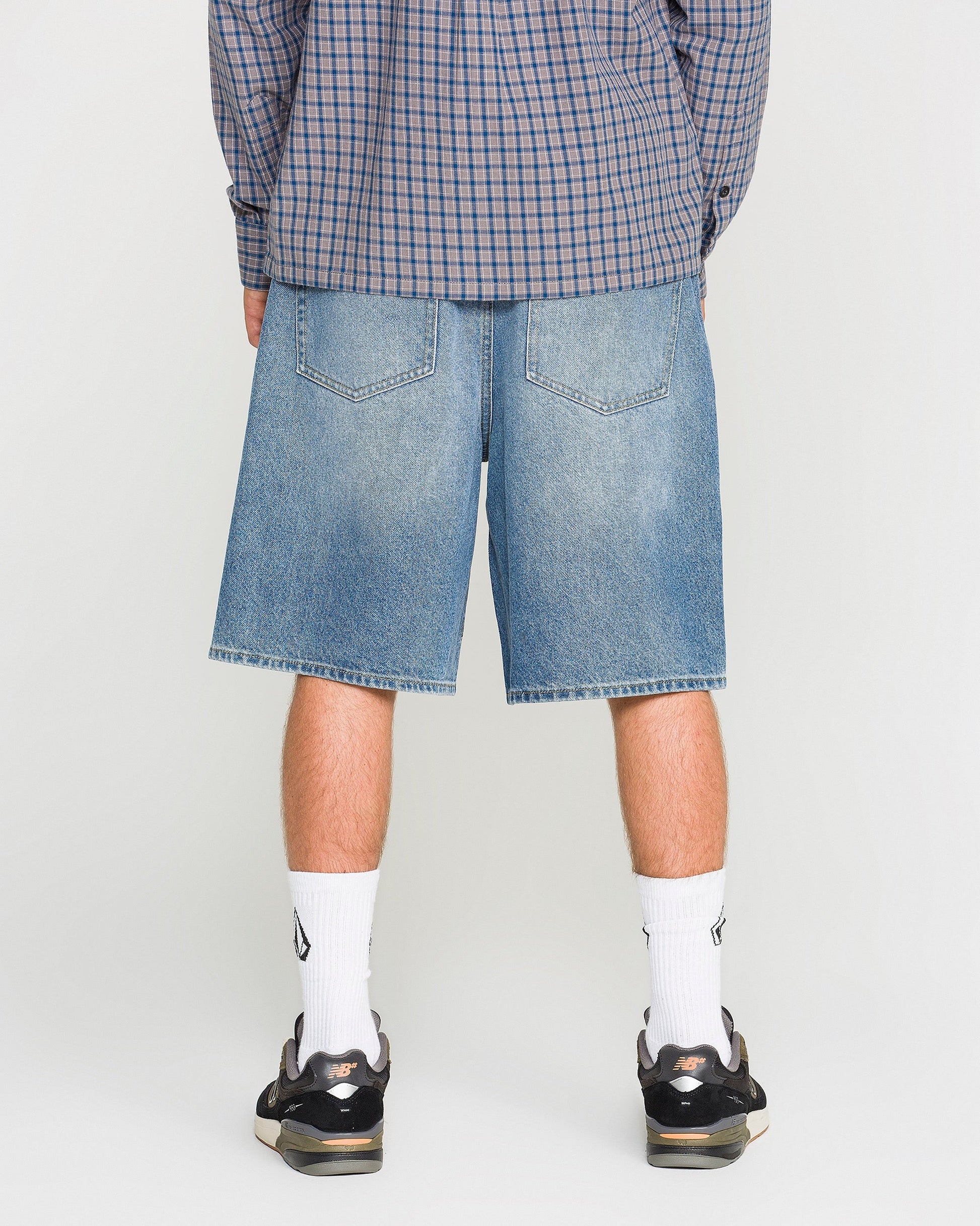 A person wearing a blue plaid shirt, Volcom Men's Billow Denim Shorts in a loose fit, white socks, and black sneakers is standing with their back to the camera against a plain background.