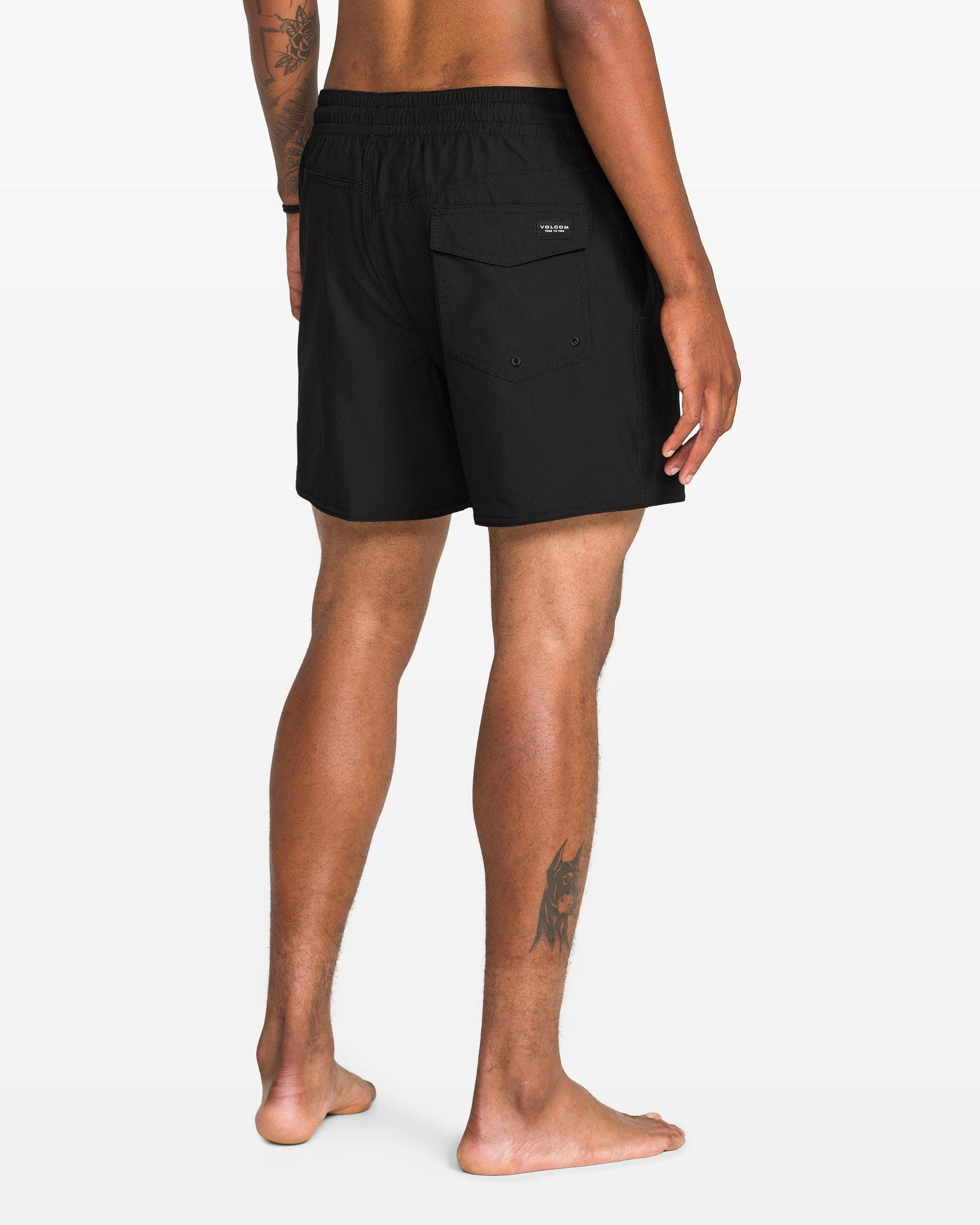 A person stands barefoot, facing away, wearing Volcom Men's Lido 2 Solid Trunks made from recycled polyester with a back pocket. Visible tattoos are on their right arm and left leg, against a plain white background.