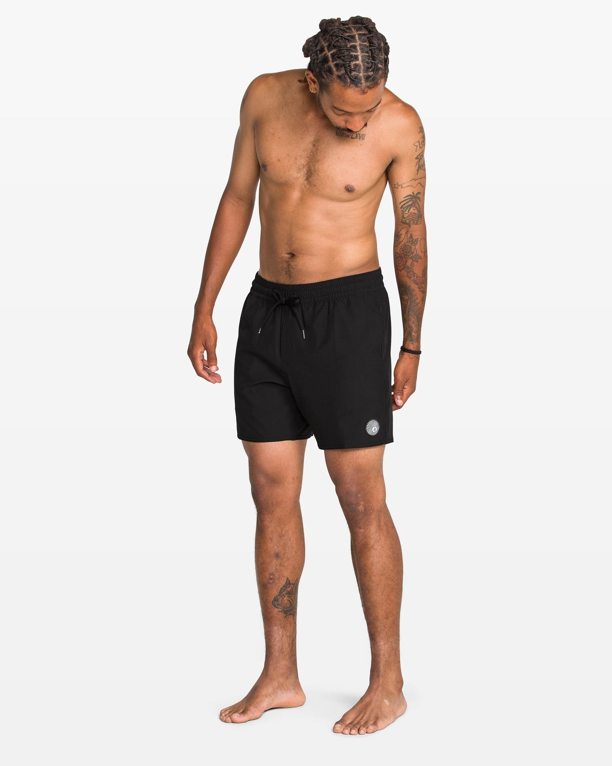 A shirtless man with braided hair and tattoos stands barefoot, looking down, wearing Volcom Men's Lido 2 Solid Trunks made from recycled polyester with mechanical stretch. The background is plain white.