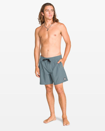 A young man with long brown hair, barefoot and wearing Volcom Men's Lido Solid 16" Trunk made from recycled polyester, stands against a plain white background, looking slightly to the side with one hand in his pocket.