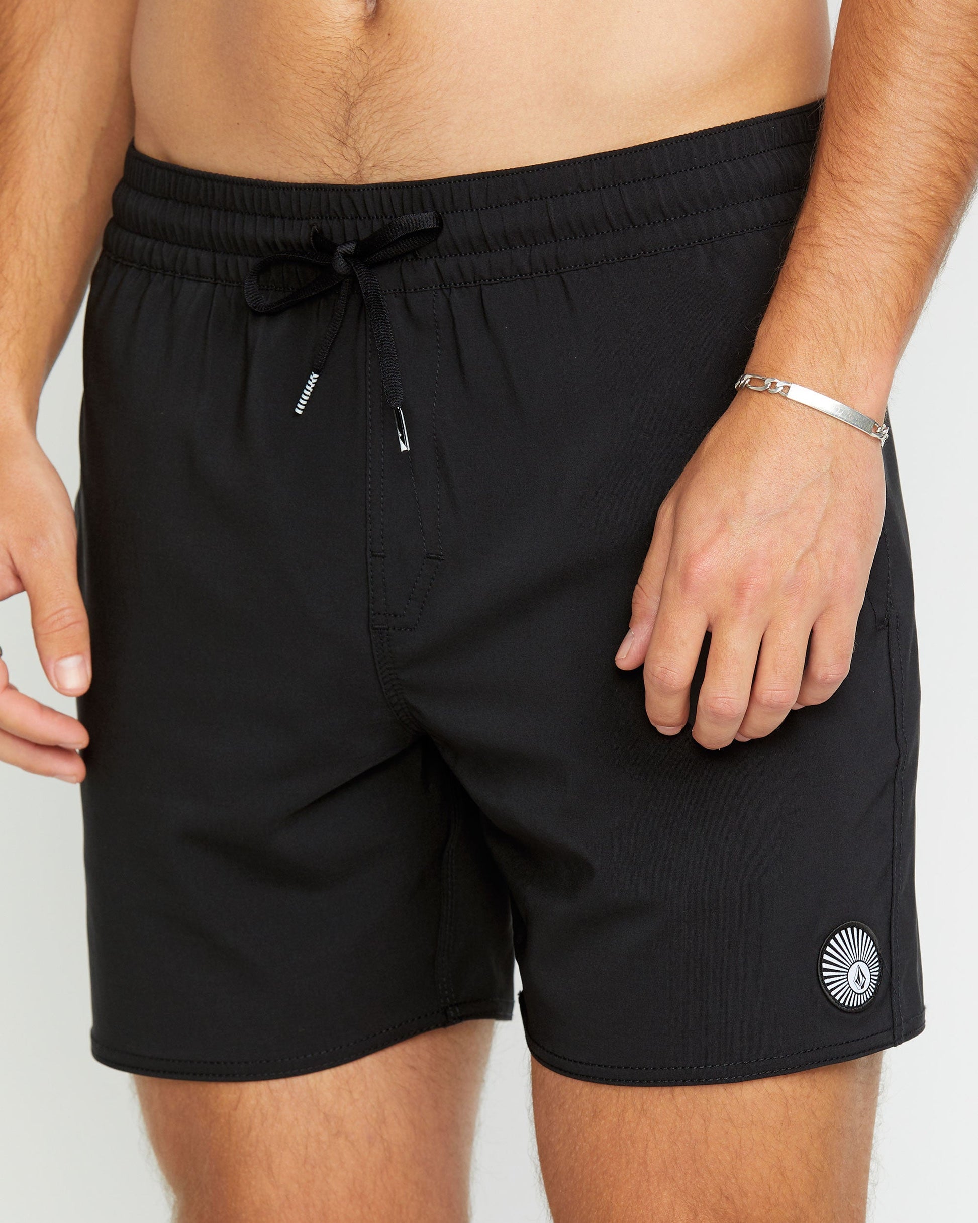 A person wears Volcom Men's Lido 2 Solid Trunks—black swim shorts made from recycled polyester, featuring a small circular white logo near the left hem, an elastic drawstring waistband, and side pockets. One hand rests relaxed by their side.