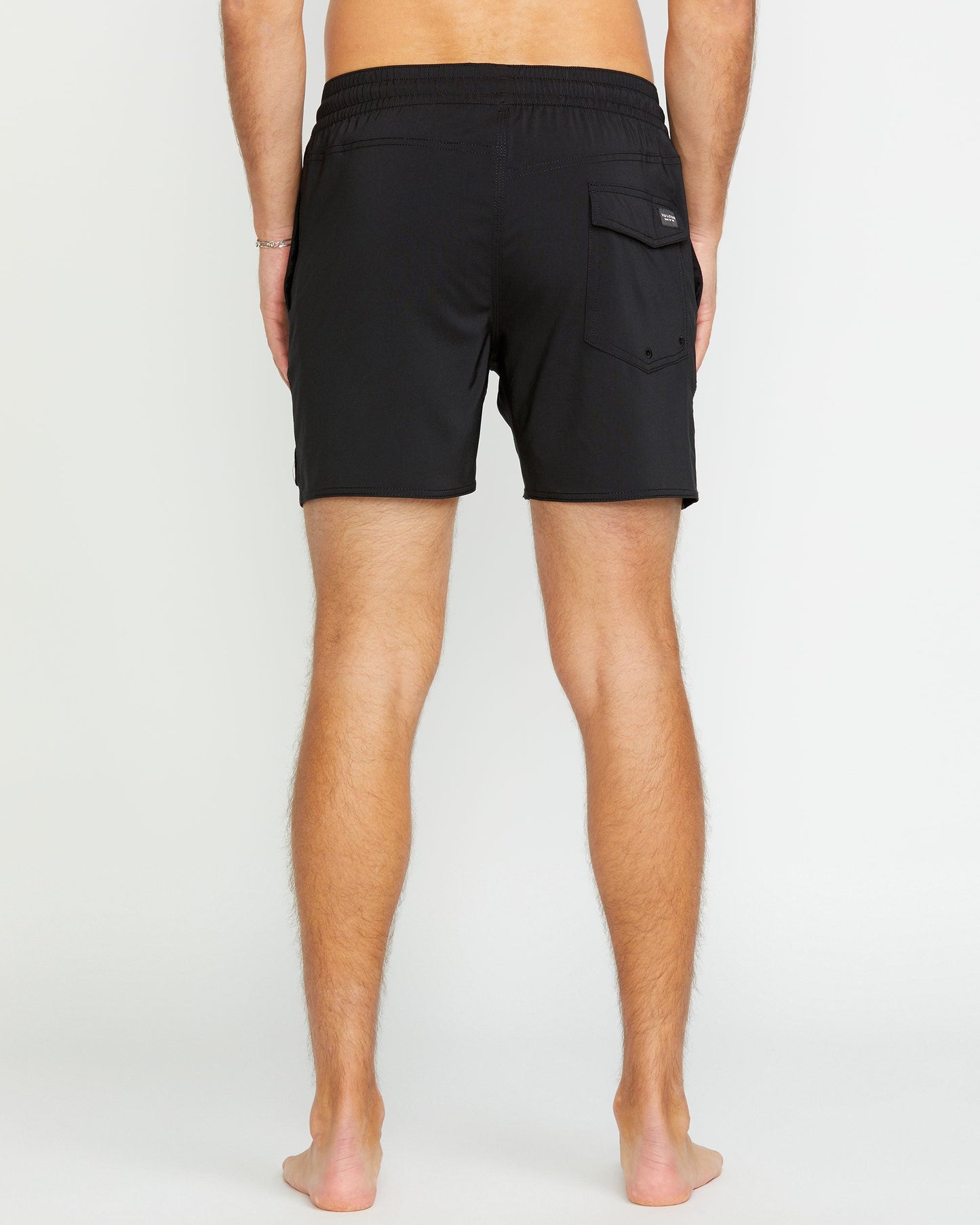 A barefoot person stands with their back to the camera, wearing Volcom Men's Lido 2 Solid Trunks—short black swim trunks made from recycled polyester, featuring an elastic waistband and a single flap pocket on the right side.