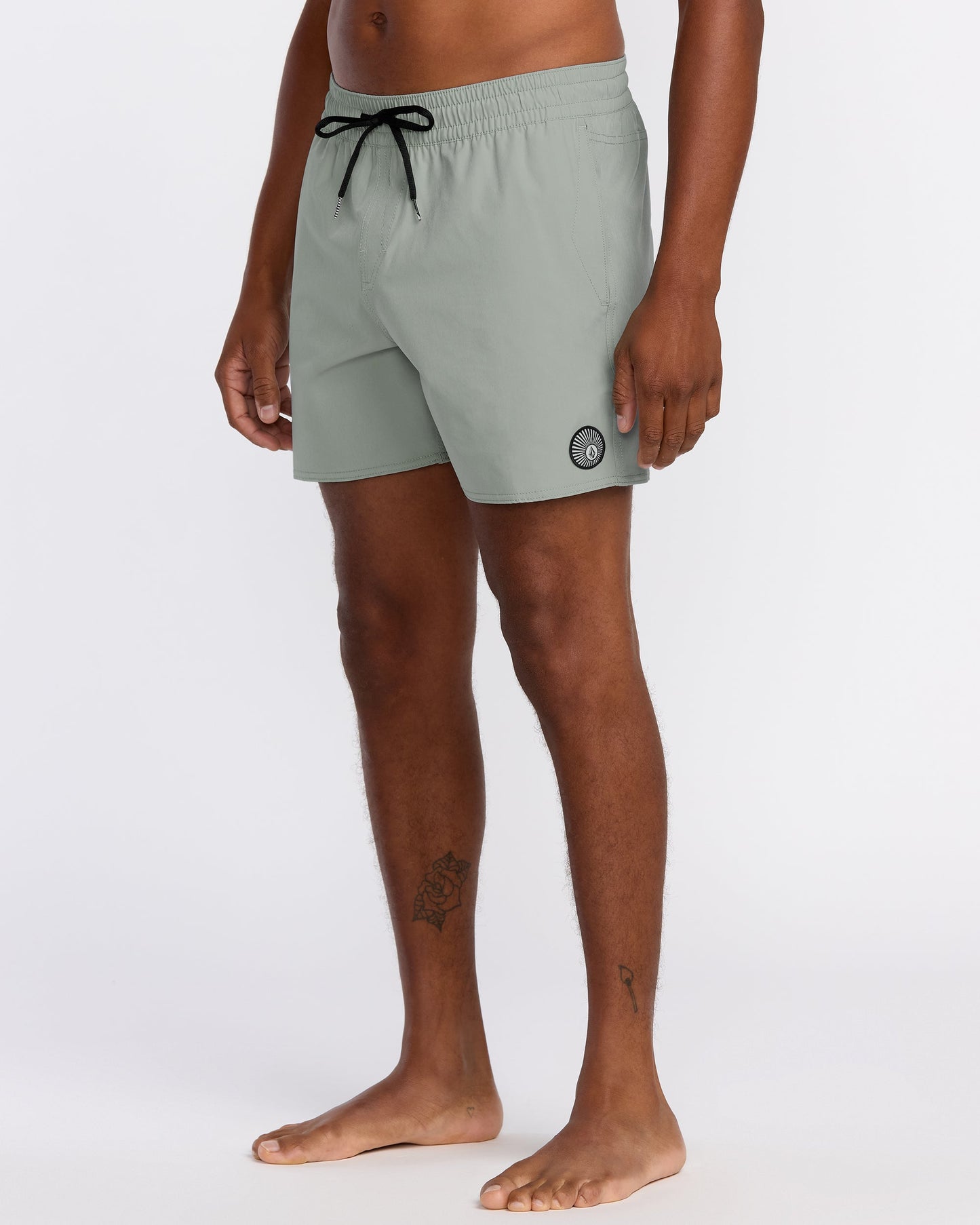 A person stands barefoot in Volcom Men's Lido Solid 16" Trunks, light gray with a black drawstring and small circular logo on the left leg. Made from recycled polyester with 2-way stretch. Torso and head not visible. Plain white background.