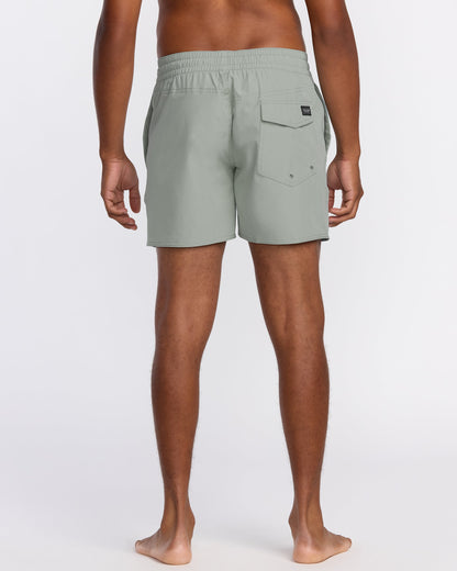 A barefoot person faces away, wearing Volcom Men's Lido Solid 16" Trunks in light gray with an elastic waistband and a right-side flap pocket, made from recycled polyester. The background is plain white.