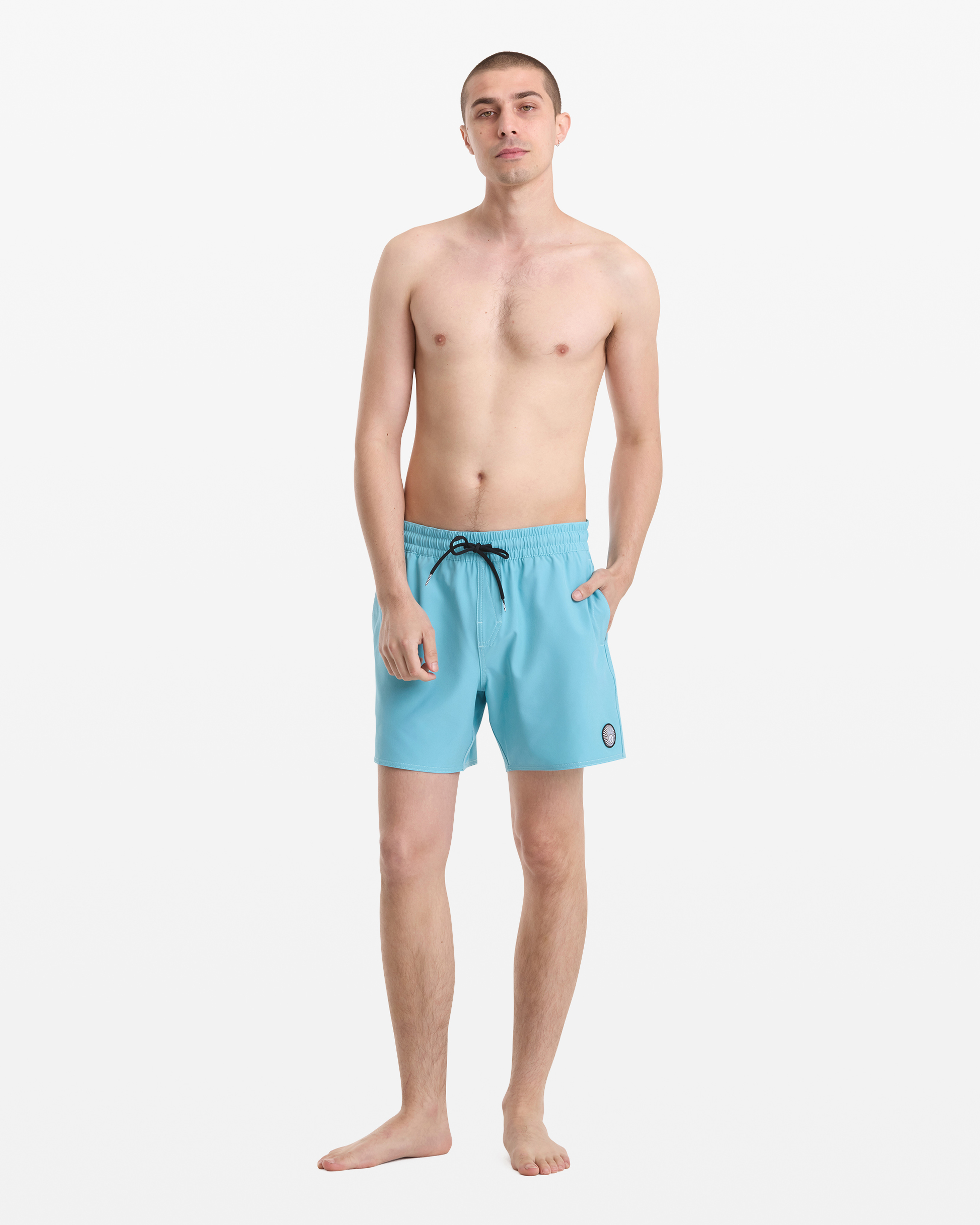 A man stands barefoot against a white background, wearing Volcom Men's Lido Solid Trunk 16 swim shorts in light blue with a black drawstring and a small round patch on the left leg. He is shirtless with short hair and his left hand in his pocket.