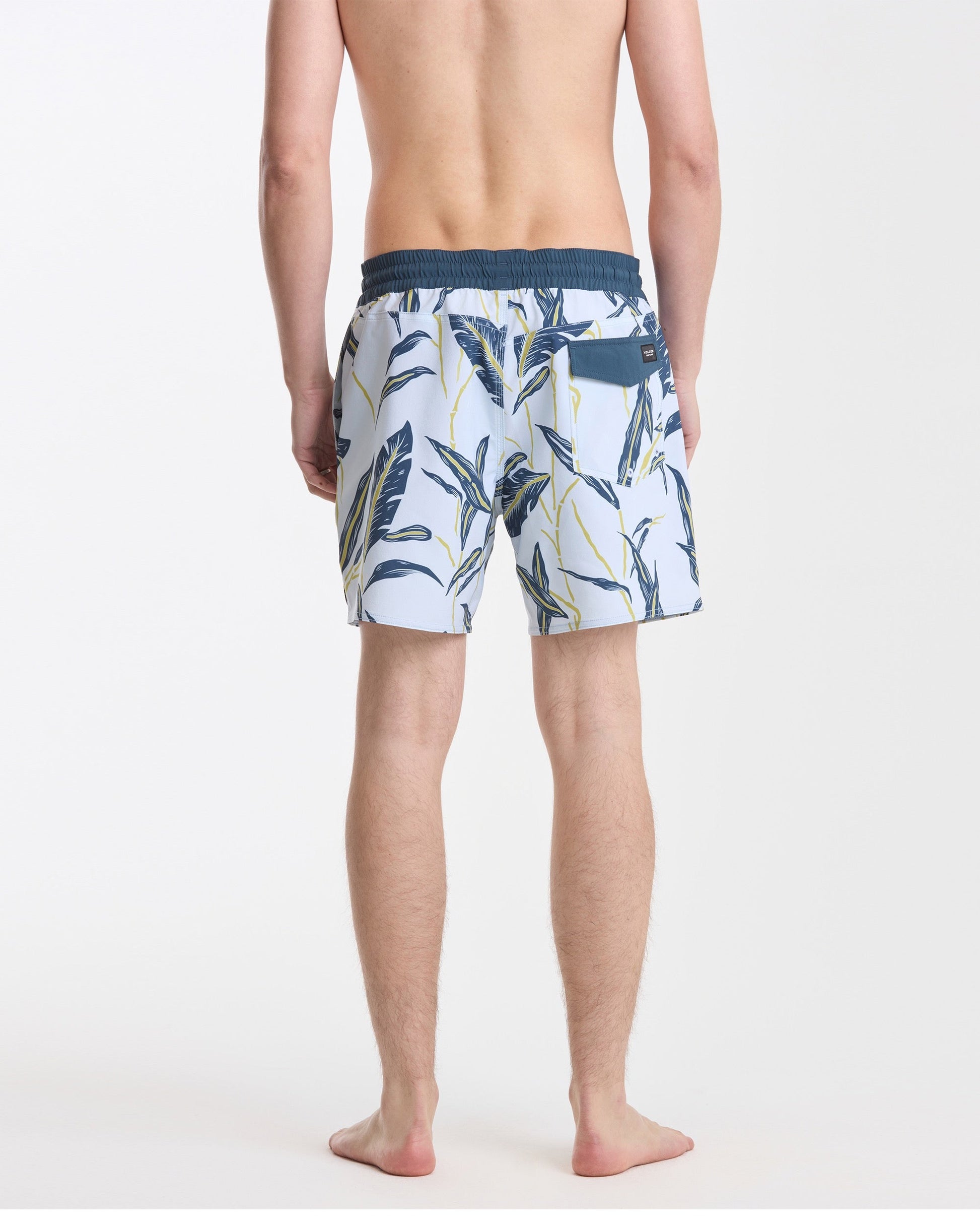 A man stands barefoot facing away in Volcom Men's Lido Print 16" Trunks, white with a navy waistband and green/yellow tropical leaf pattern, made from stretchy polyester-elastane. Background is plain white.
