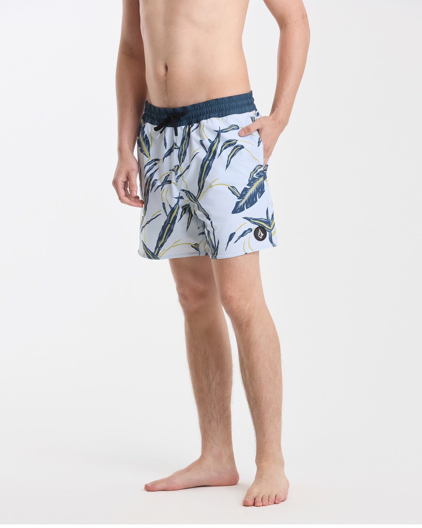 A man stands barefoot against a white background wearing Volcom’s Men's Lido Print 16" Trunks—light blue with a dark blue and yellow tropical leaf pattern, stretch polyester-elastane, dark blue waistband, back flap pocket, left hand in pocket.