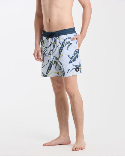 A man stands barefoot against a white background wearing Volcom’s Men's Lido Print 16" Trunks—light blue with a dark blue and yellow tropical leaf pattern, stretch polyester-elastane, dark blue waistband, back flap pocket, left hand in pocket.