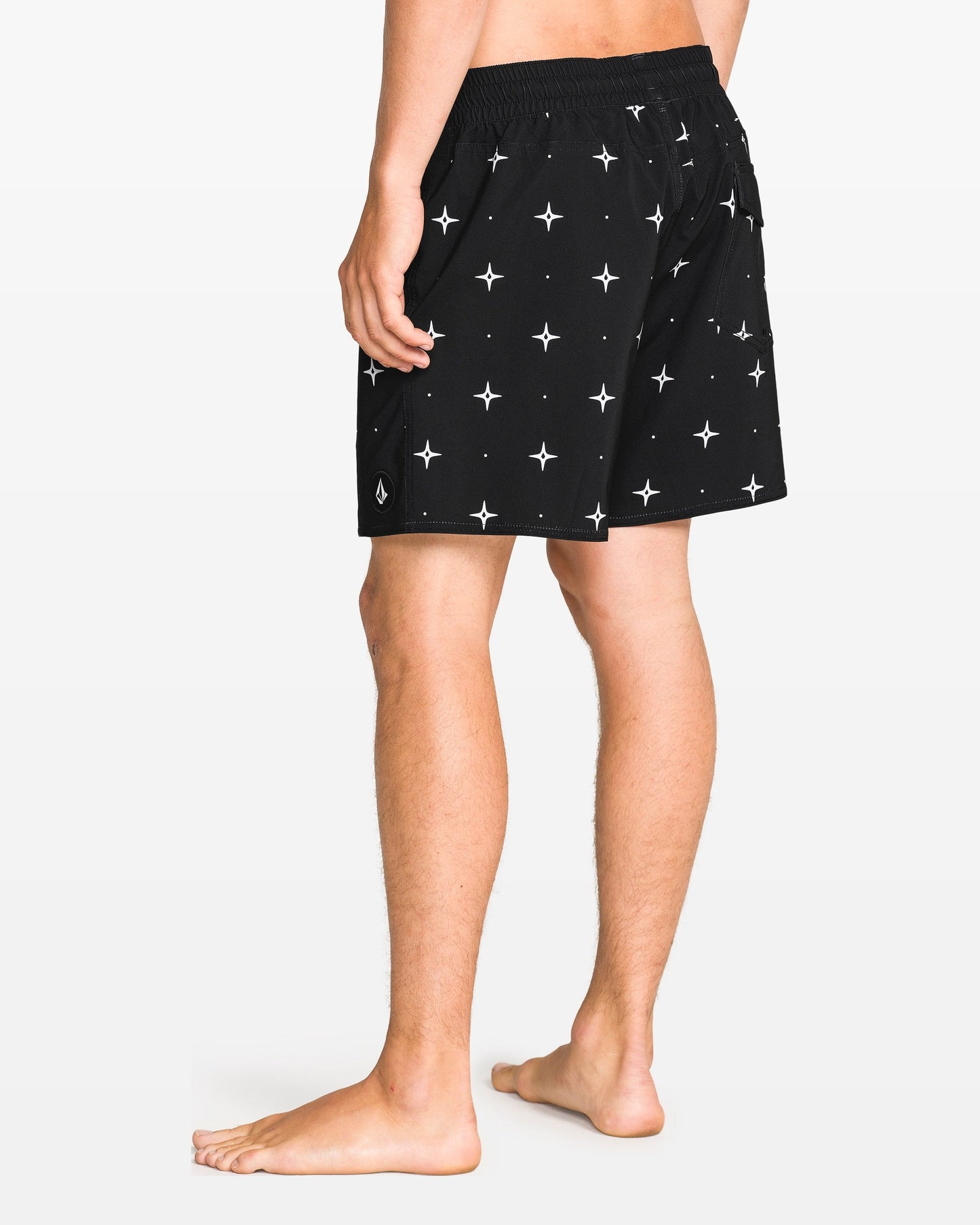 A barefoot person, shown from the waist down and facing away, wears Volcom Men's Lido Print 16" Trunk shorts in black with a white star pattern and 4-way stretch. The background is solid white.