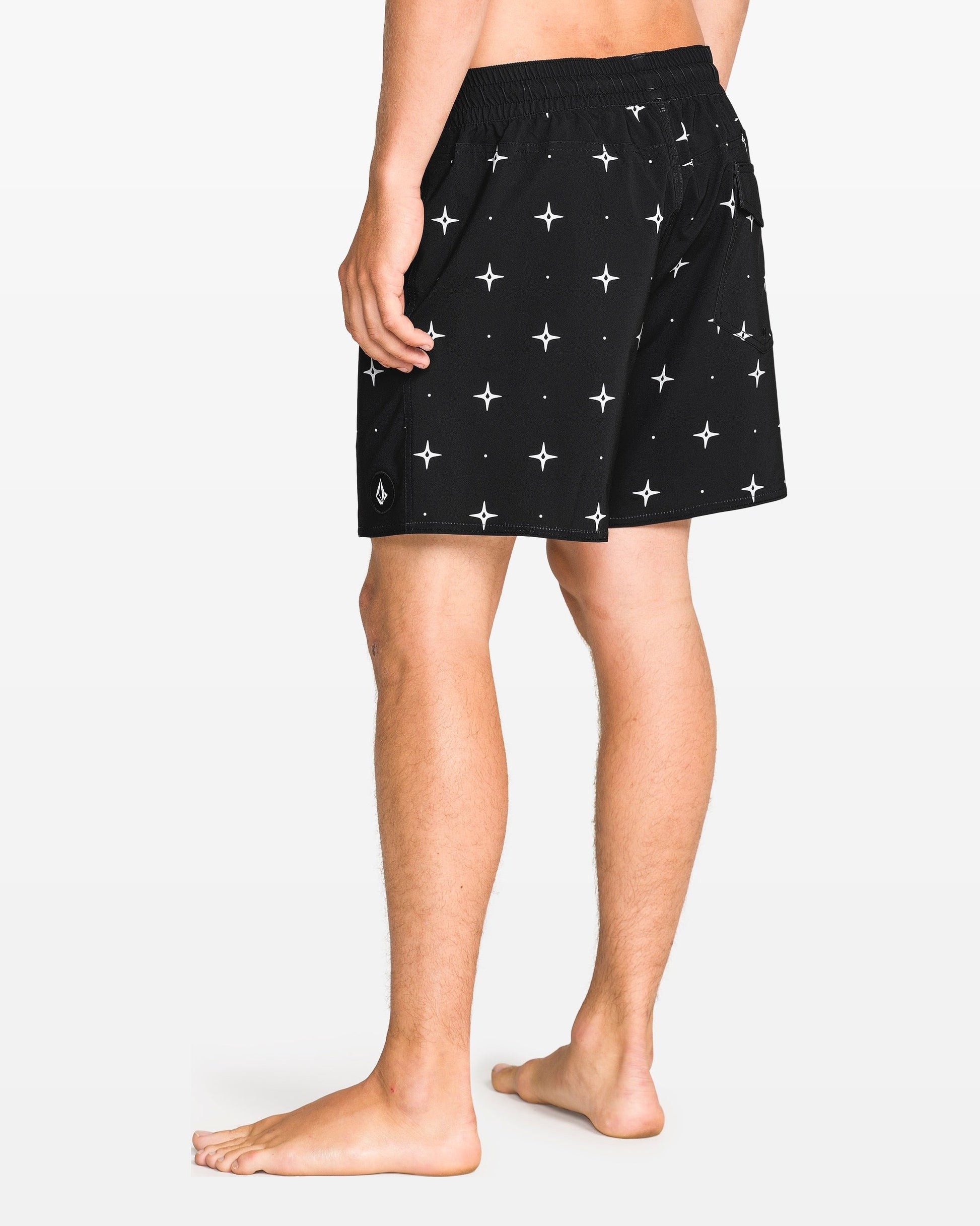 A barefoot person, shown from the waist down and facing away, wears Volcom Men's Lido Print 16" Trunk shorts in black with a white star pattern and 4-way stretch. The background is solid white.